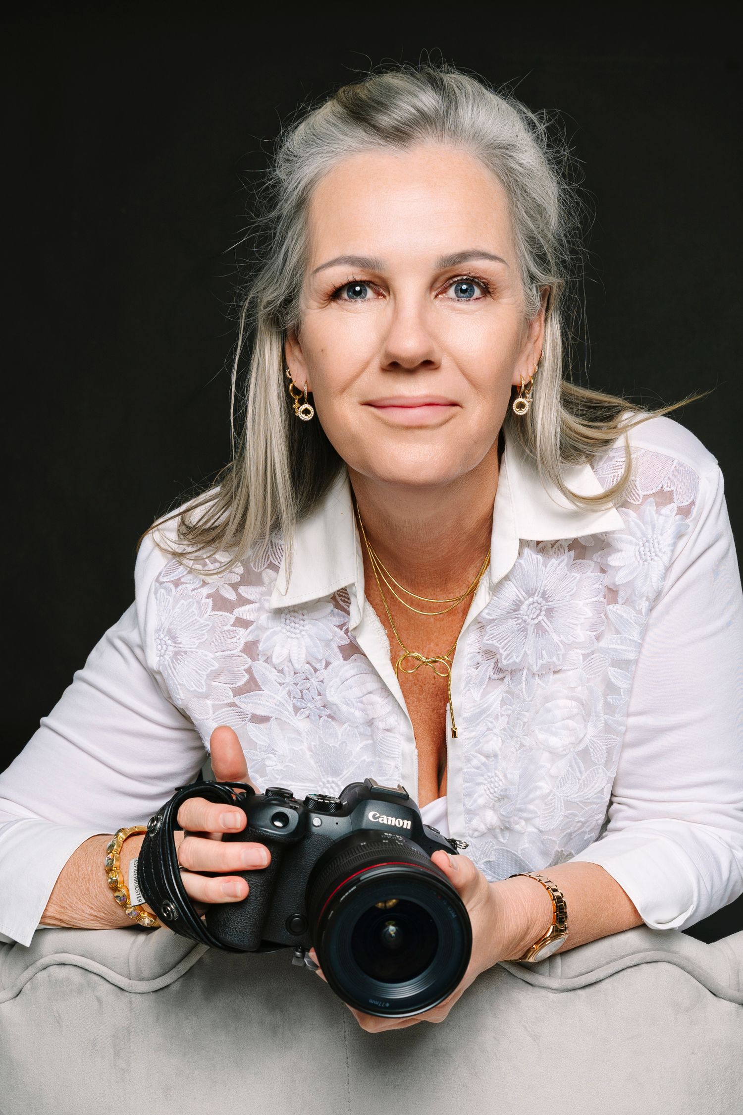 Middle-aged woman with gray hair holding a Canon camera, wearing a white blouse and gold jewelry. Wollongong photographer.