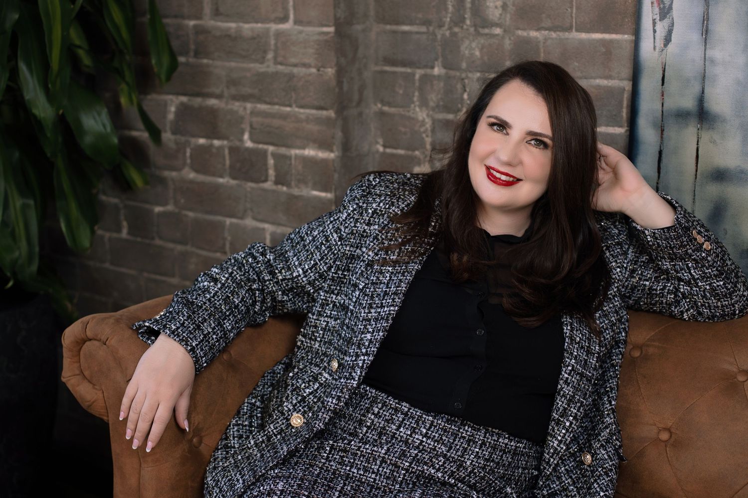 Woman in tweed blazer smiling on leather sofa against brick wall. Wollongong personal branding photography.