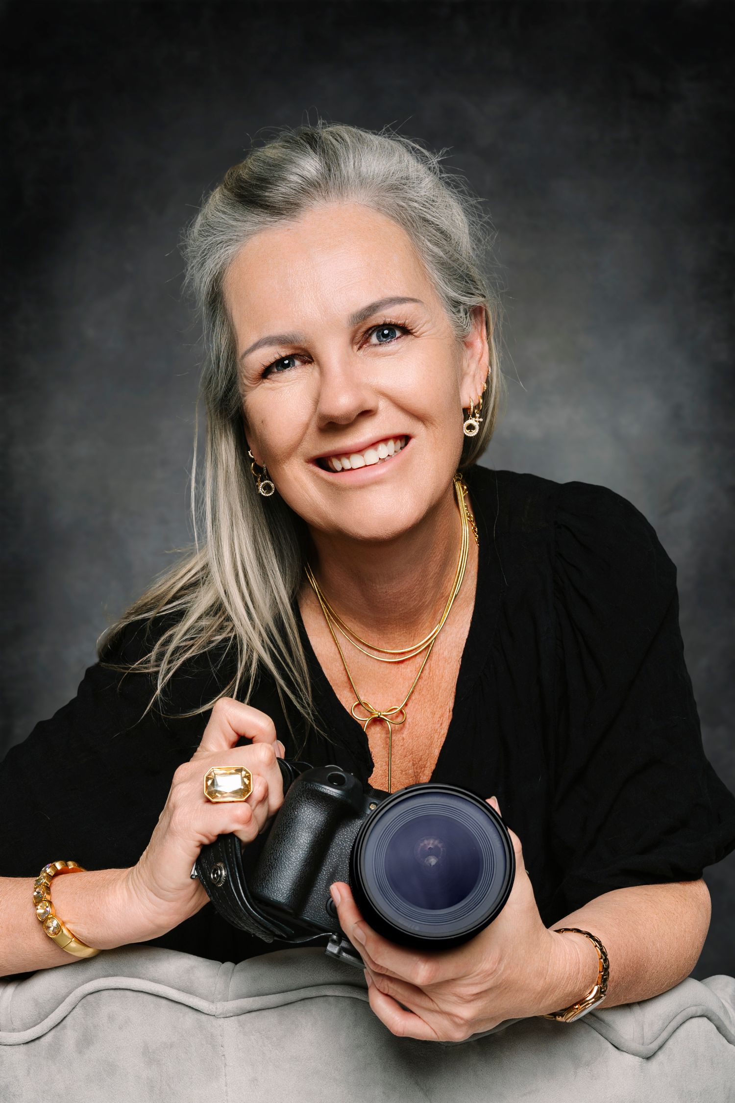 Smiling woman with gray hair holding a professional camera, wearing gold jewelry. Portrait photography. Wollongong photographer