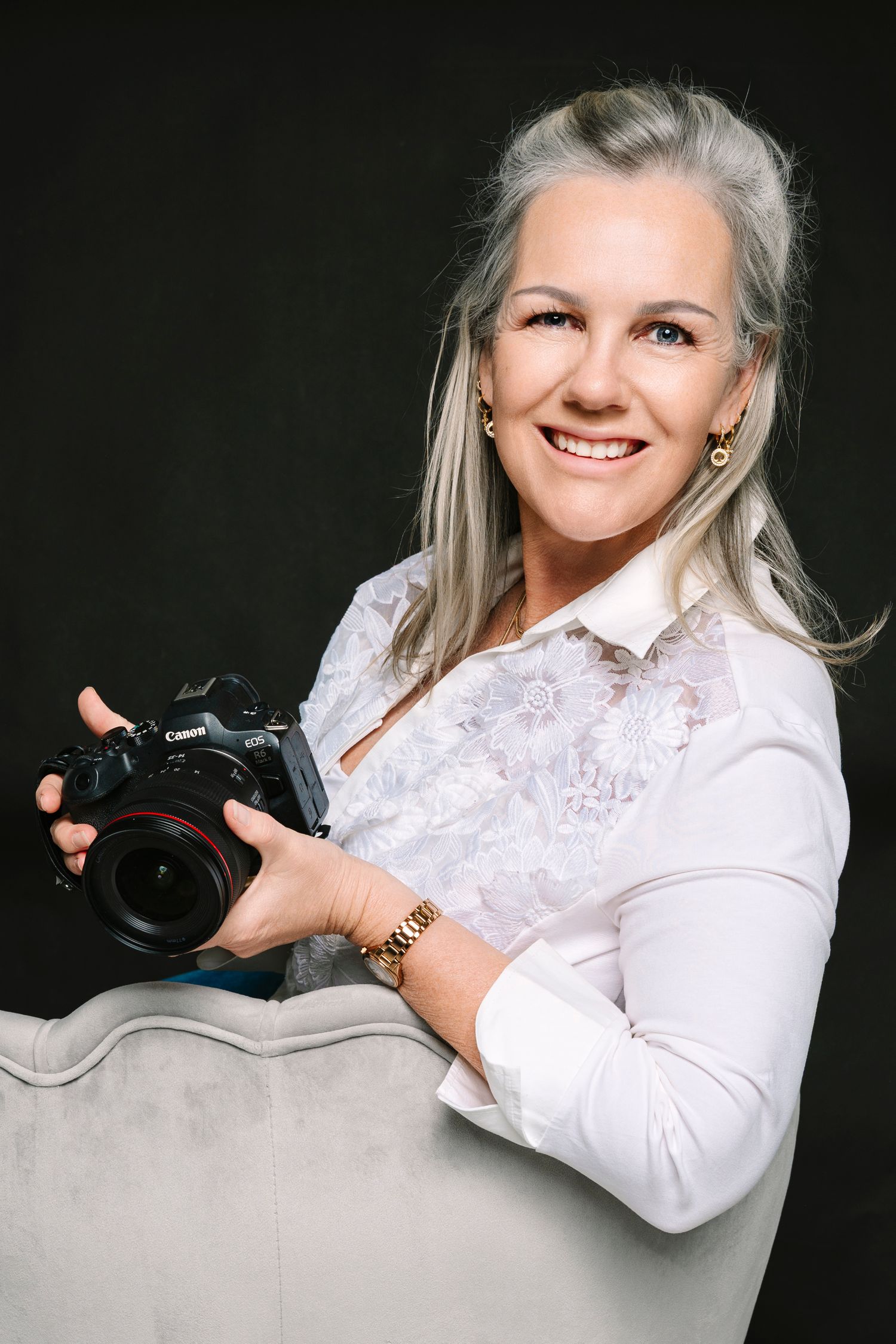 Smiling woman with gray hair holding a Canon camera, wearing a white blouse against dark background. Wollongong photographer