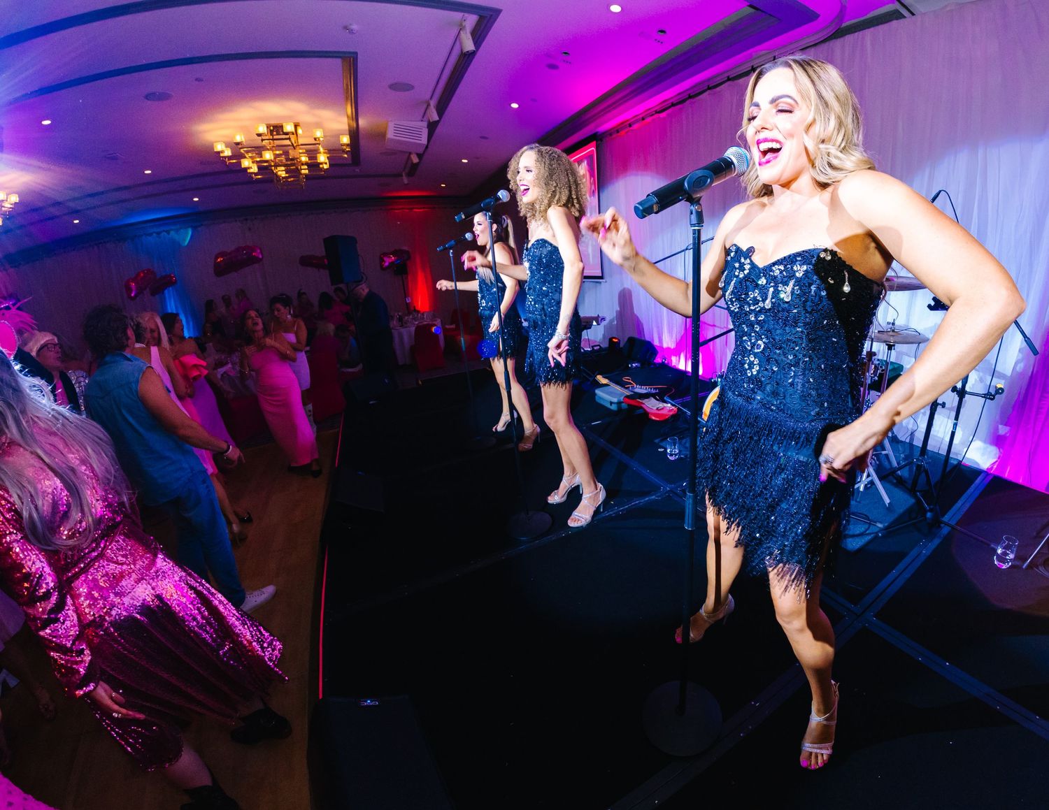 Wollongong Corporate Event Photographer. Female performers in sequined dresses sing and dance on stage at an event with pink lighting.
