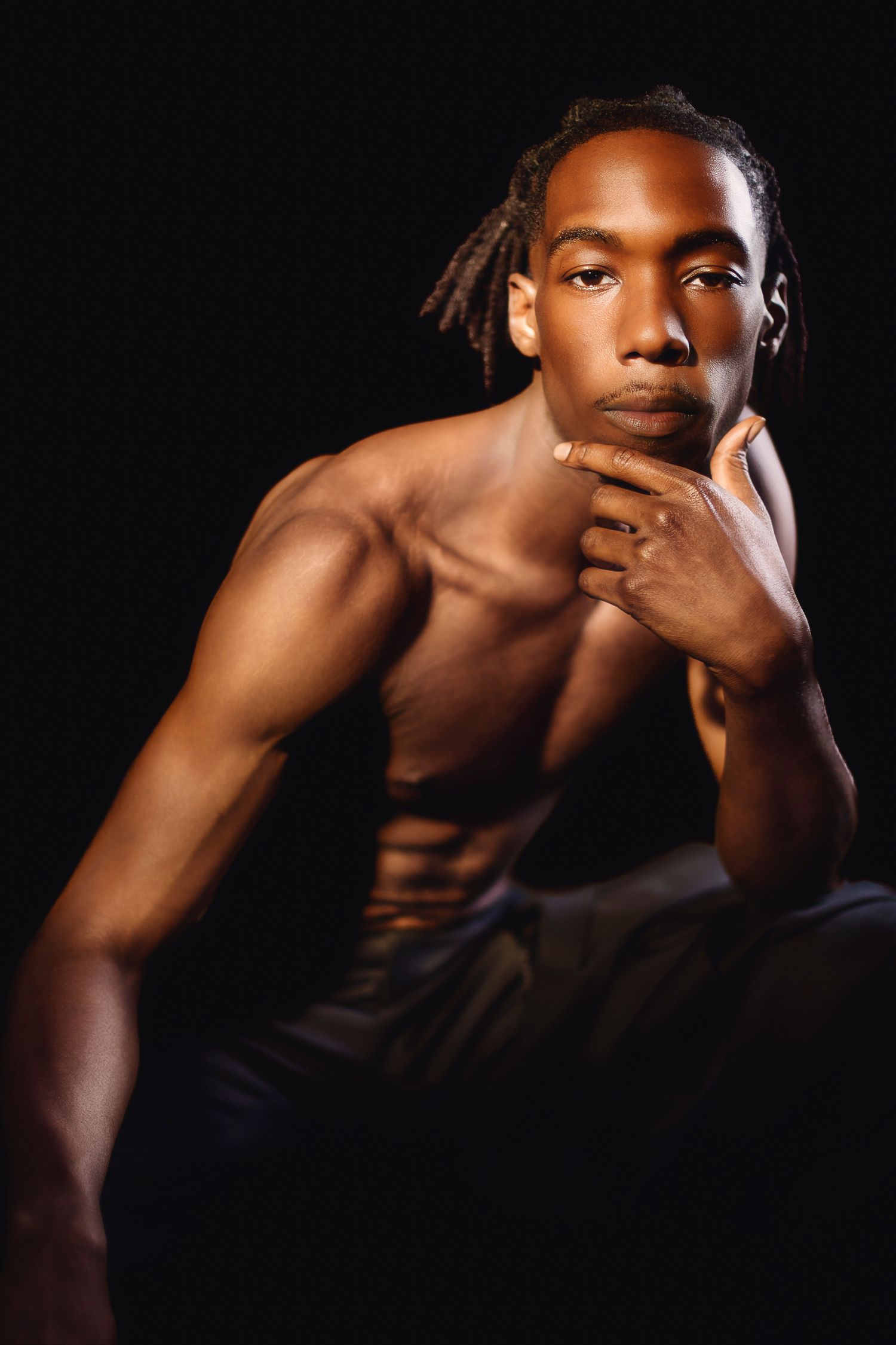 Wollongong Portrait Photographer. Muscular shirtless Black man with dreadlocks posing thoughtfully against dark background.