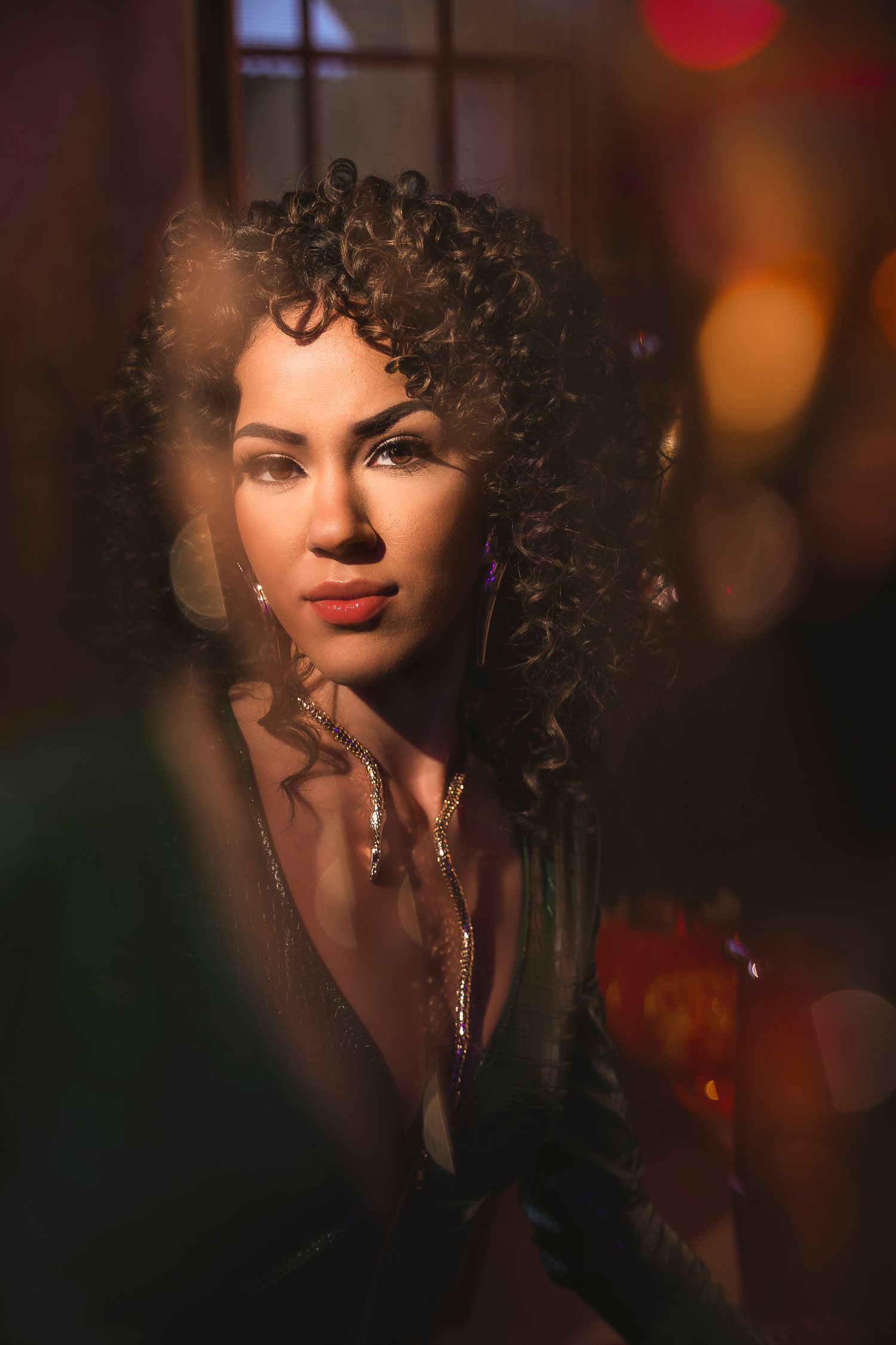 Wollongong Portrait Photographer. A woman with curly hair and gold necklaces poses in warm bokeh lighting, wearing a deep V-neck top.