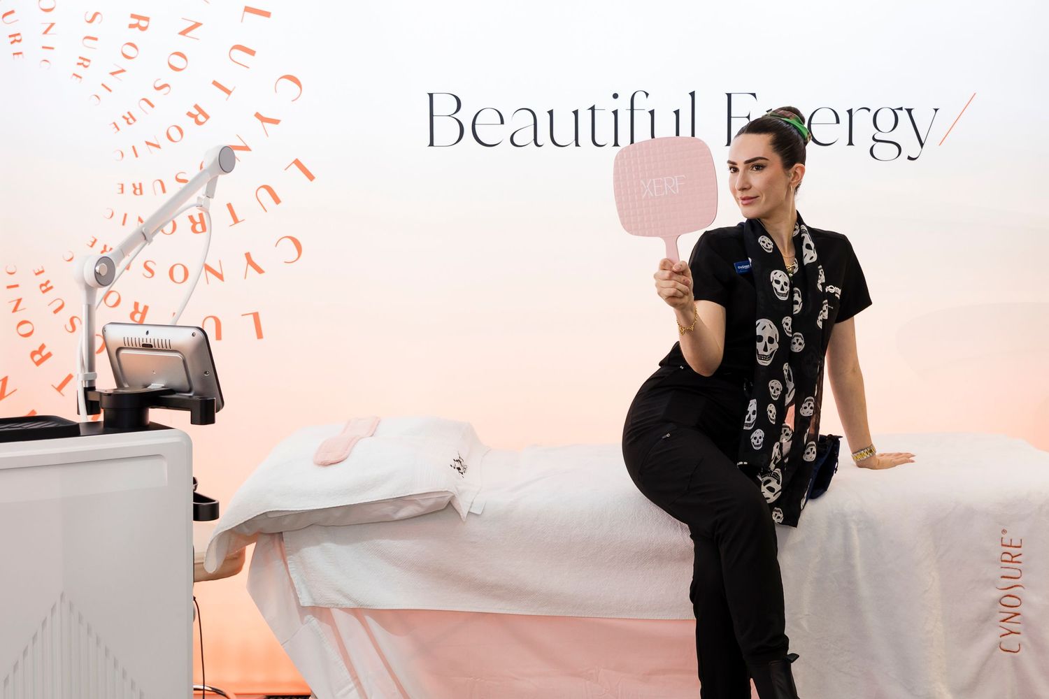 A woman in black floral scrubs holds a pink mirror at a Beautiful Energy spa treatment event. Wollongong Corporate Branding Photographer