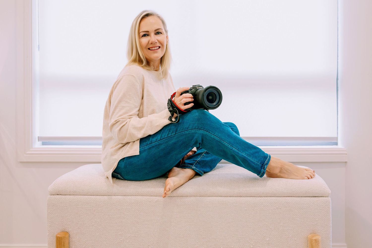 Wollongong Photographer. Smiling blonde woman sitting on ottoman holding Canon camera in bright white room wearing jeans and beige sweater.