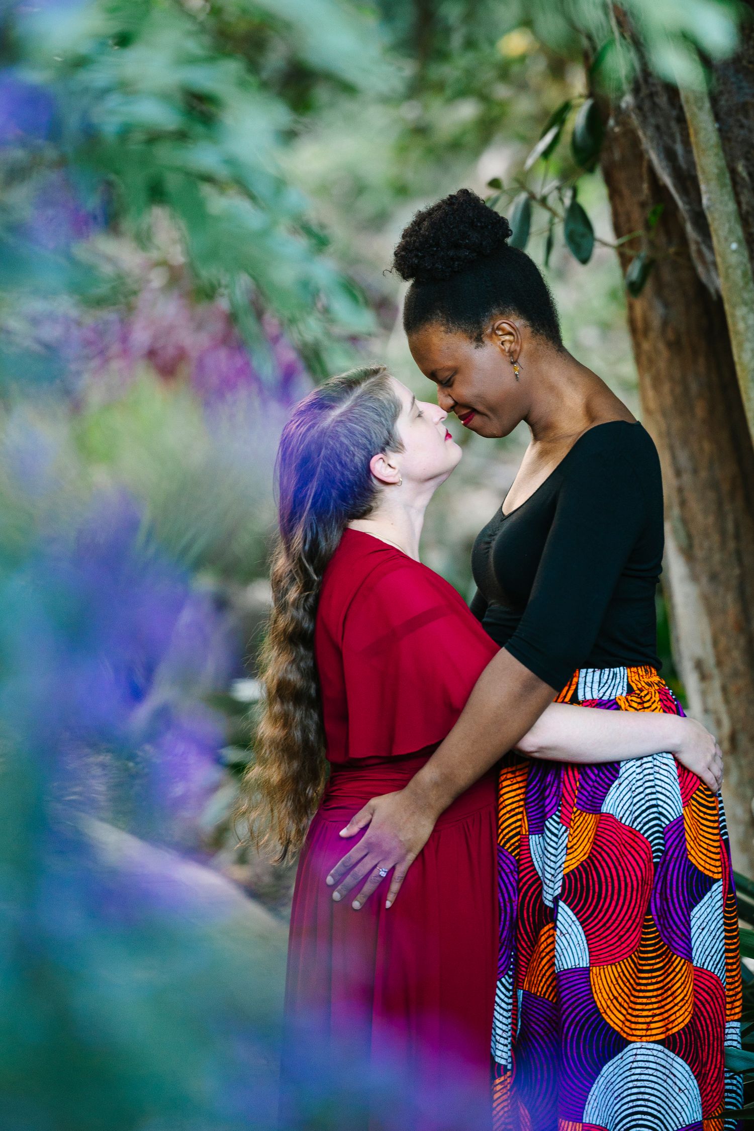 Two women in colorful dresses share a romantic moment in a lush garden with purple and green foliage. Wollongong Engagement Photographer
