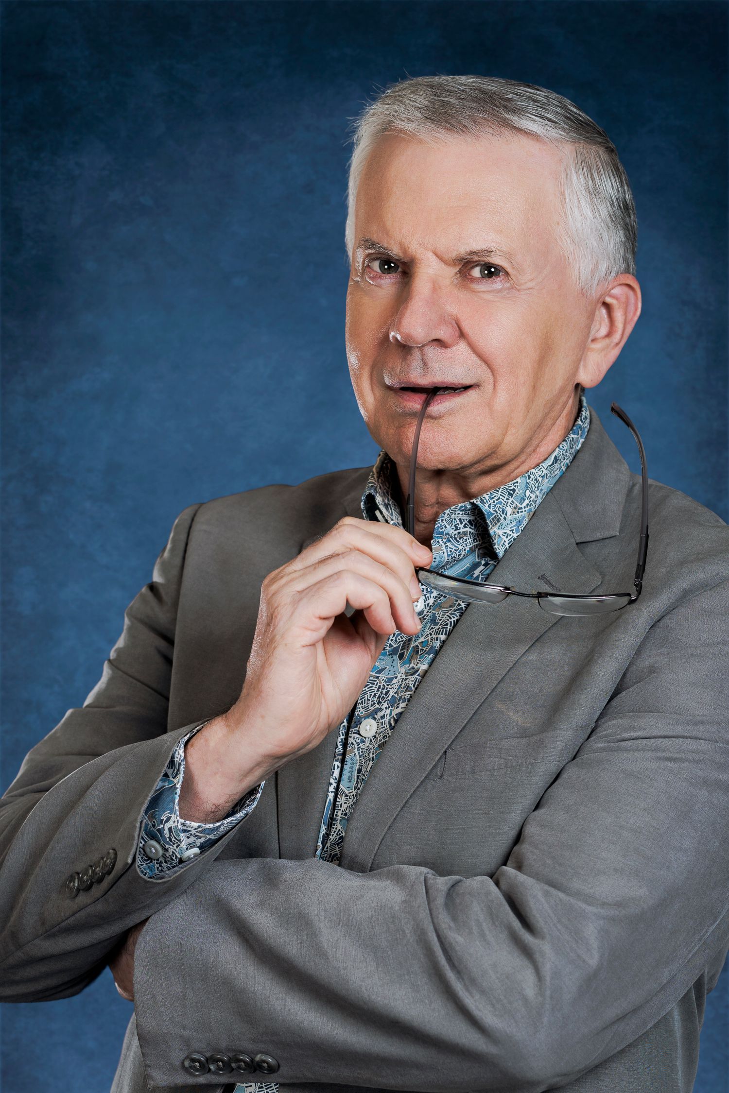 Elderly man in gray blazer holding glasses thoughtfully, posing against a blue studio background. Wollongong Headshot Photographer