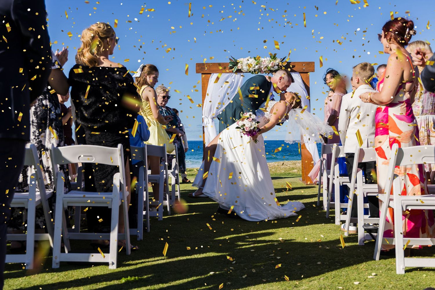 Bride and groom share first kiss  with confetti falling on green lawn near ocean. Wollongong Wedding Photographer
