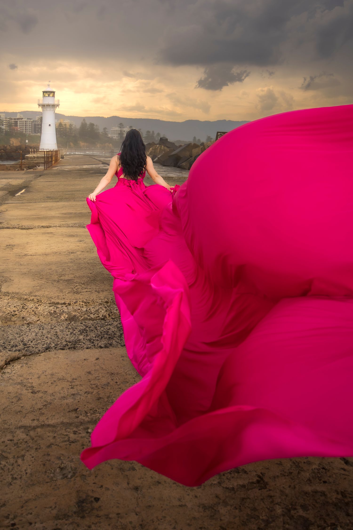 Woman in flowing pink gown walks toward lighthouse at dramatic sunset. Wollongong Flying Dress Photographer