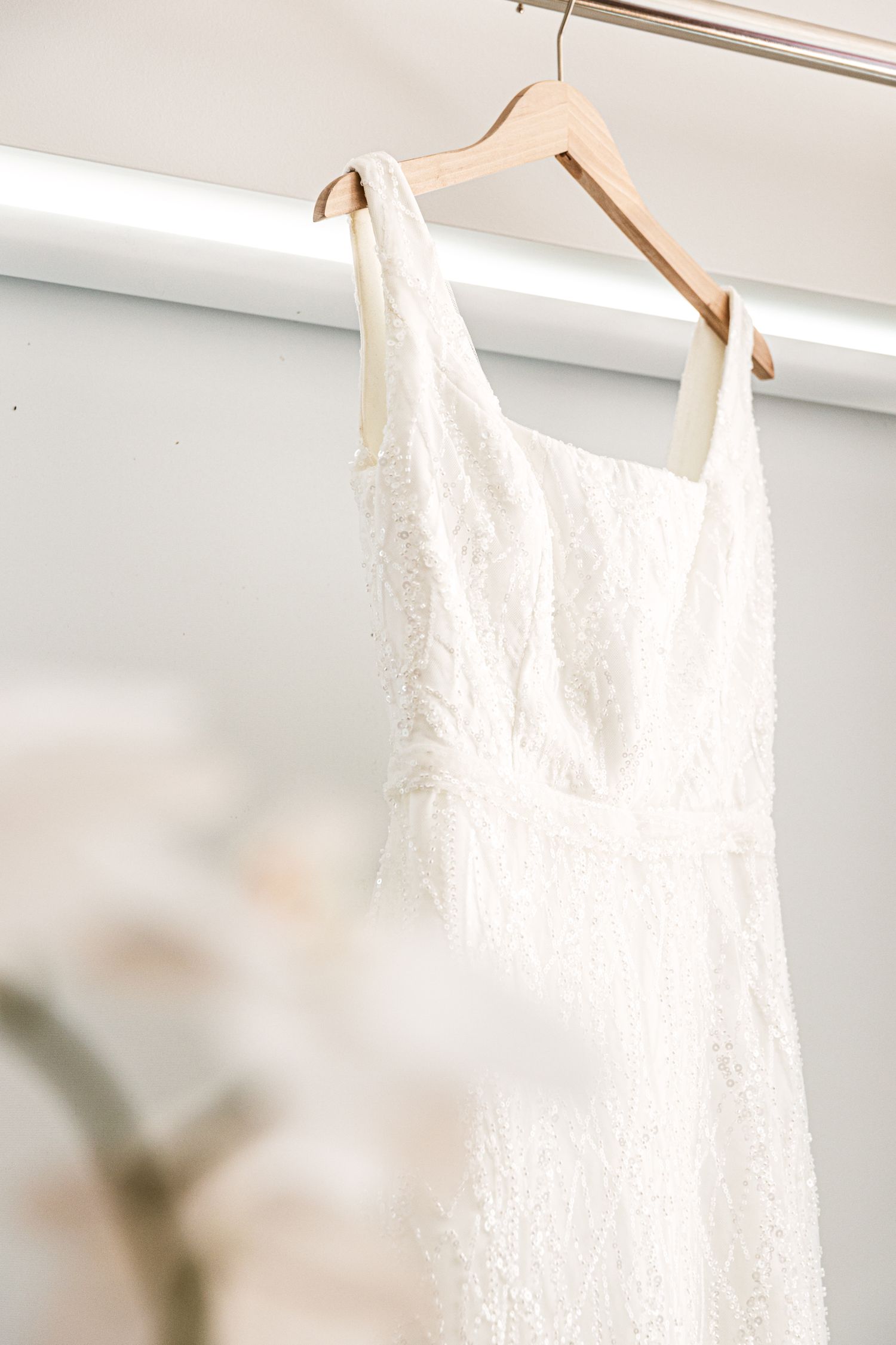 White lace wedding dress hanging on a wooden hanger against a soft white background. Wollongong Wedding Photographer