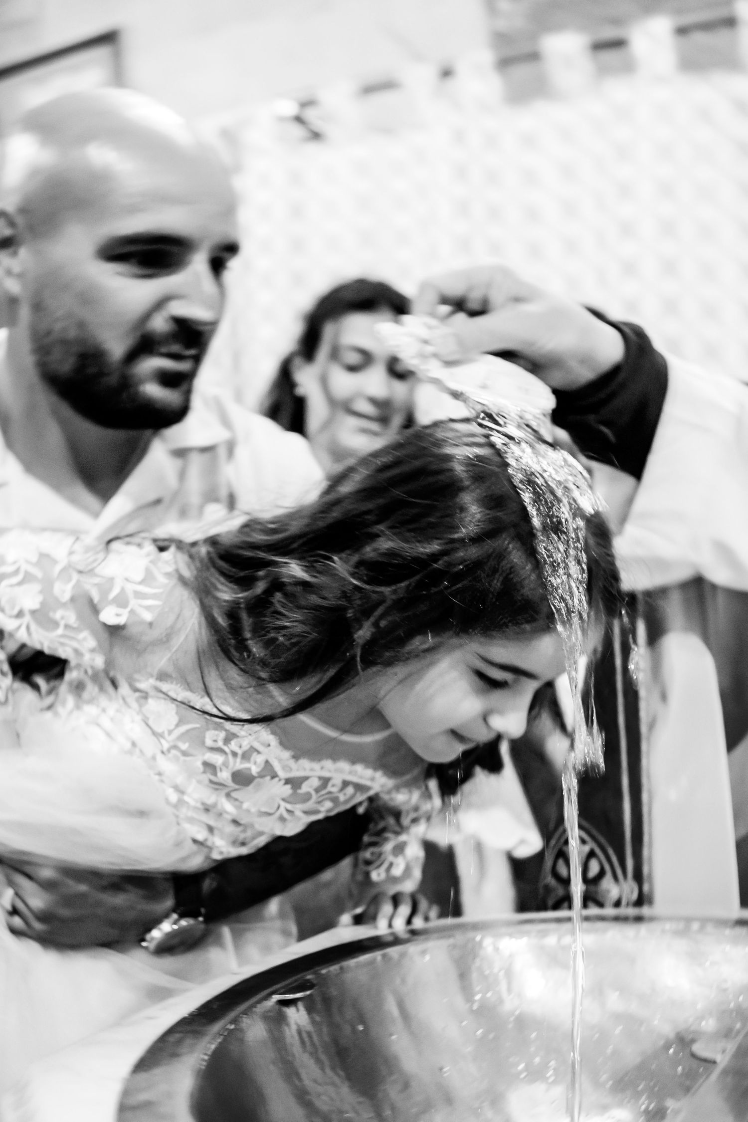 Black and white photo of a girl being baptized, water poured over her head into a metal basin by a bald man.
