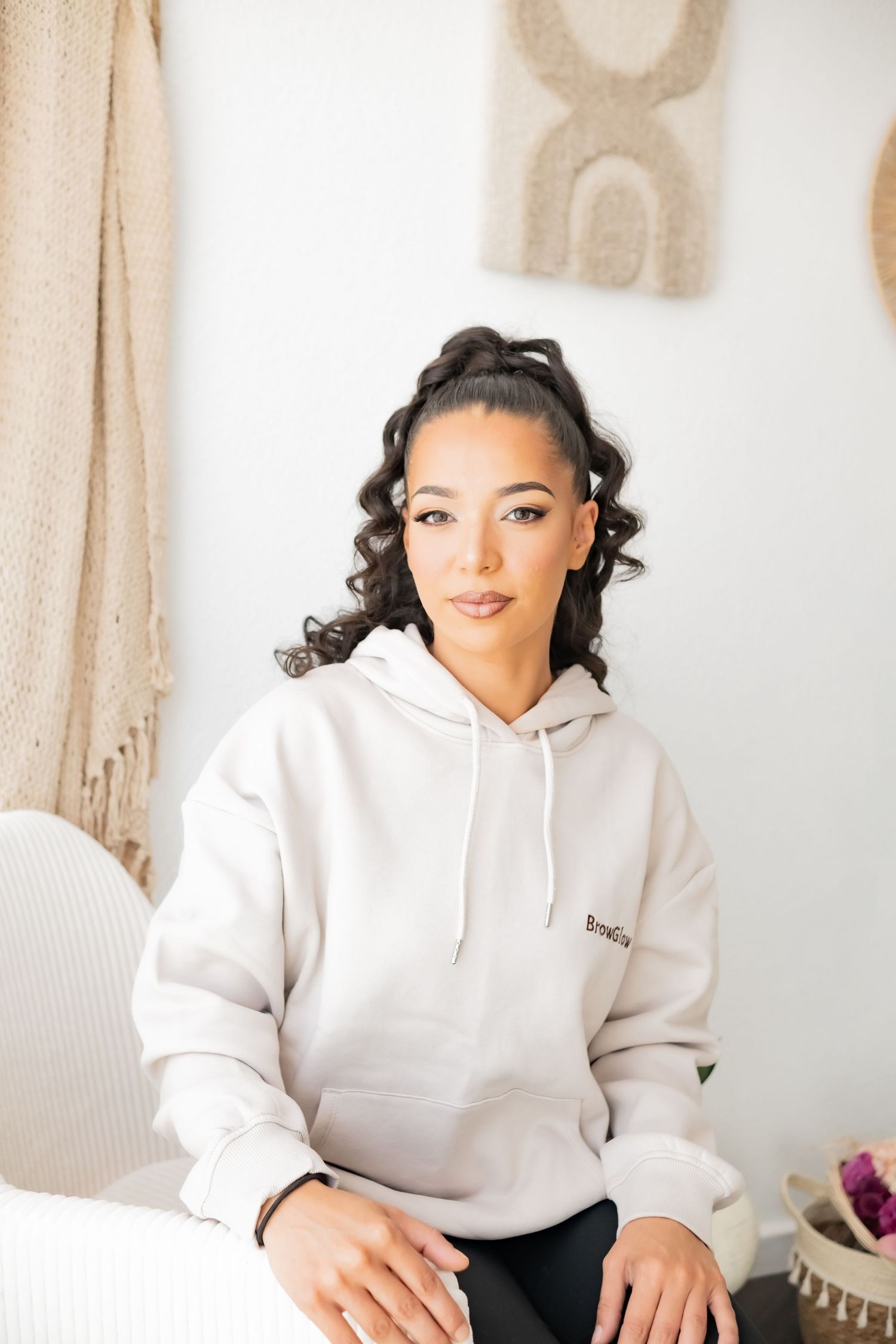 Young woman with curly hair wearing a beige hoodie sits confidently in a bright, bohemian-styled room.