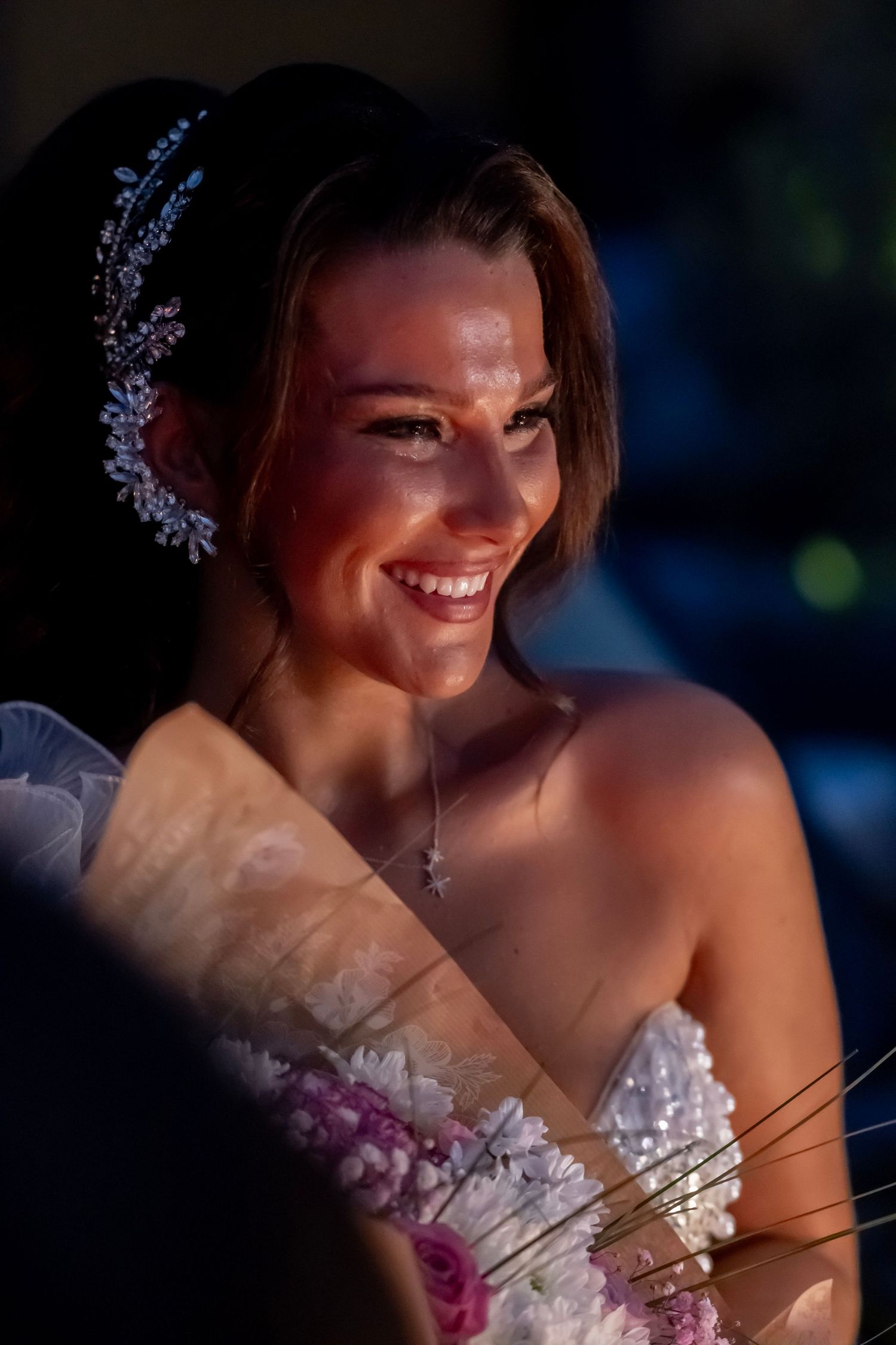 A glowing bride smiles radiantly in a white wedding dress with delicate floral details and decorative hair accessories.
