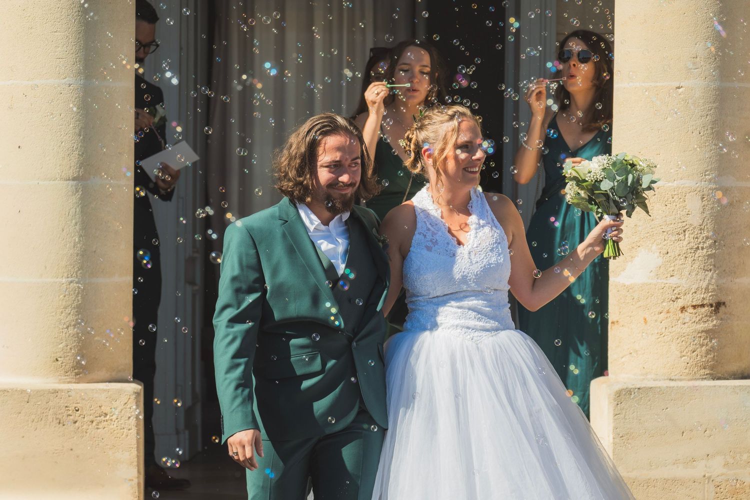 Joyful newlywed couple exits church surrounded by bubbles, bride in white gown holding bouquet, groom in green jacket.