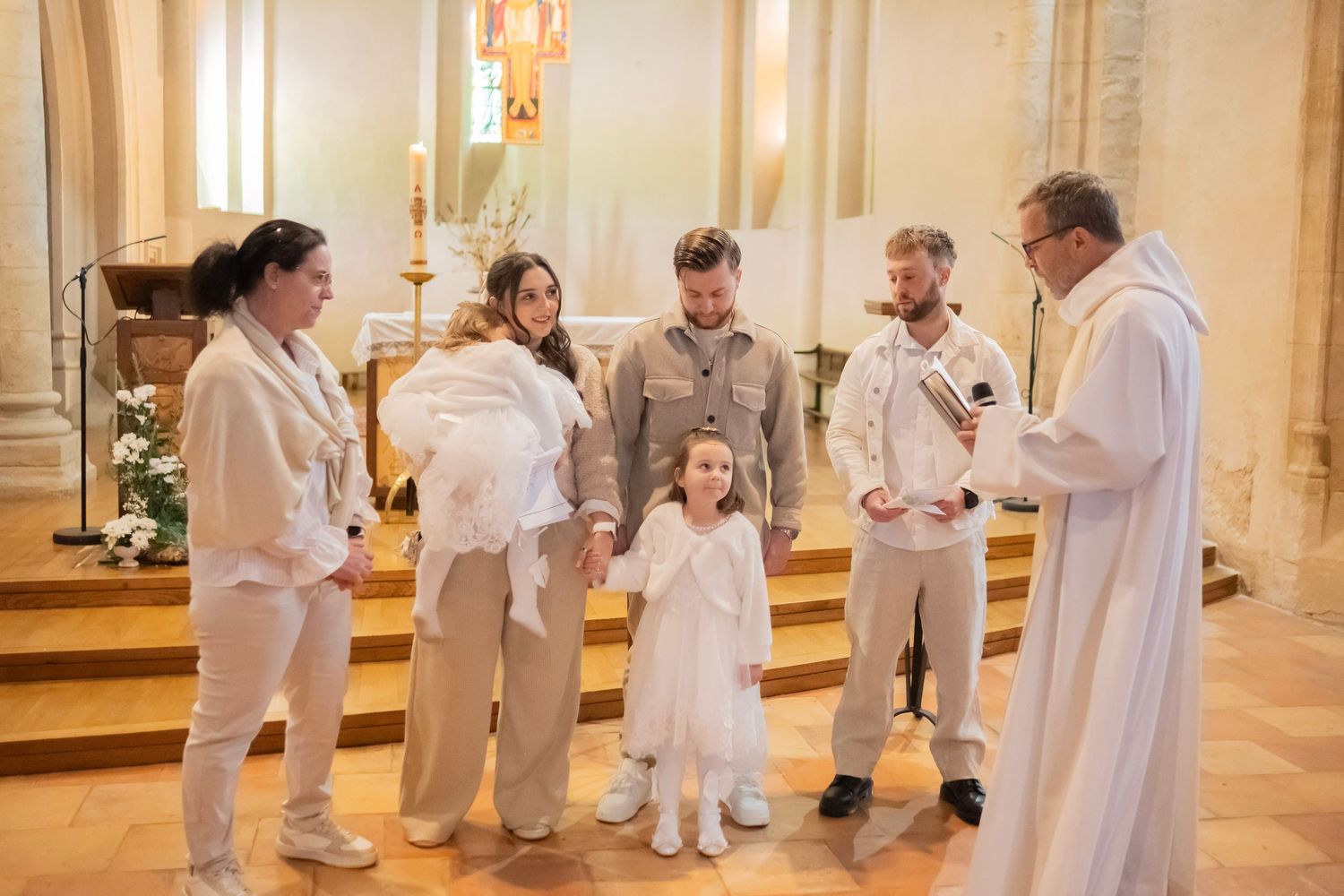 A baptismal ceremony taking place in a church with people dressed in white and beige standing at the altar with a priest.