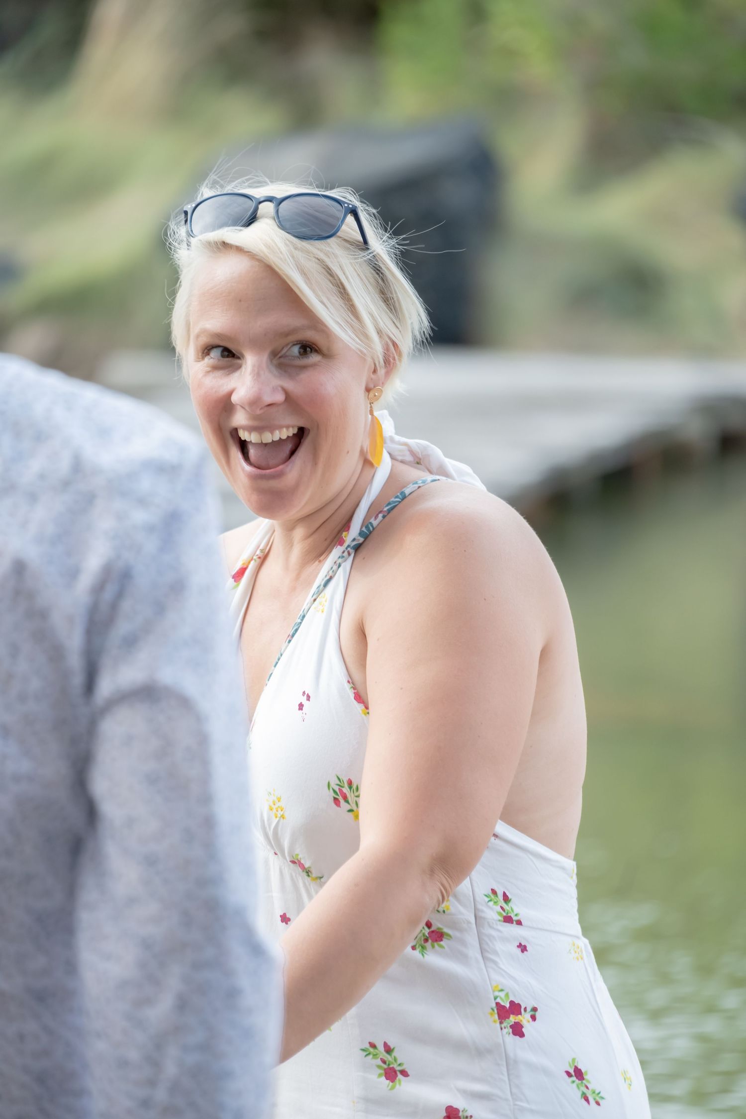 A joyful person wearing a white floral sundress and sunglasses laughs while enjoying a summer day by the water.