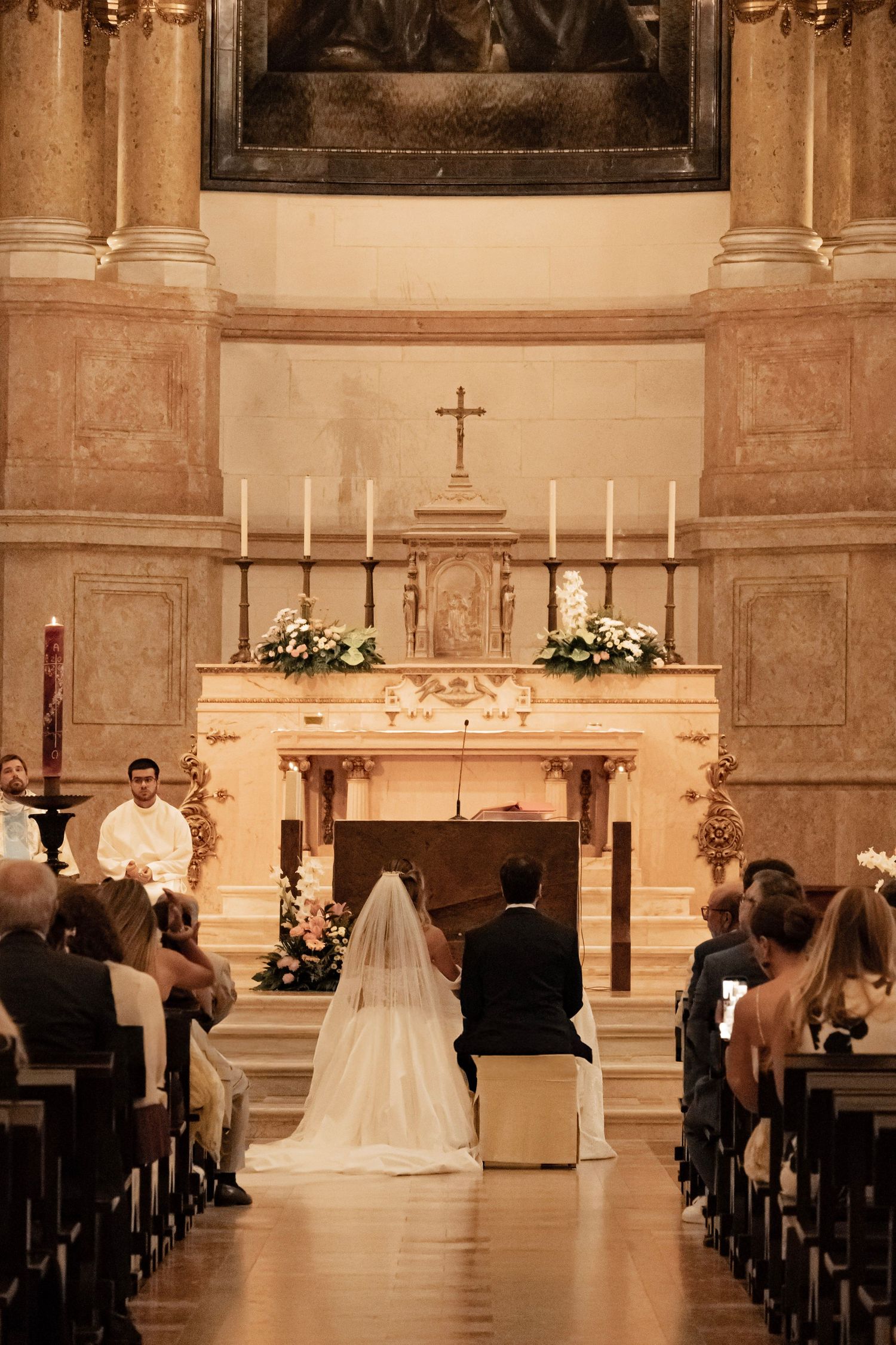 Bride and groom kneel at ornate church altar during Catholic wedding ceremony with candles and floral arrangements.