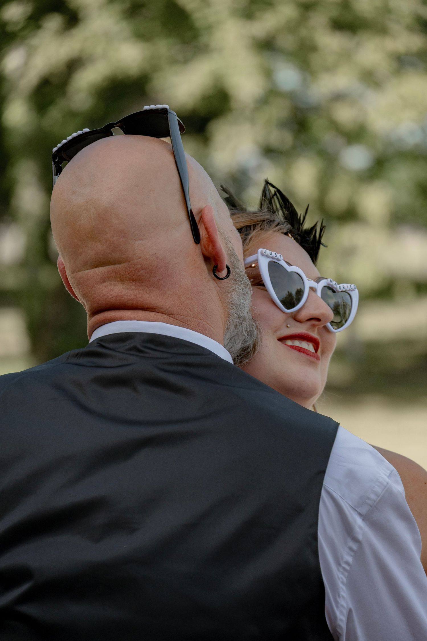 A couple embracing outdoors, woman in heart-shaped white sunglasses smiling, man in black vest with shaved head.