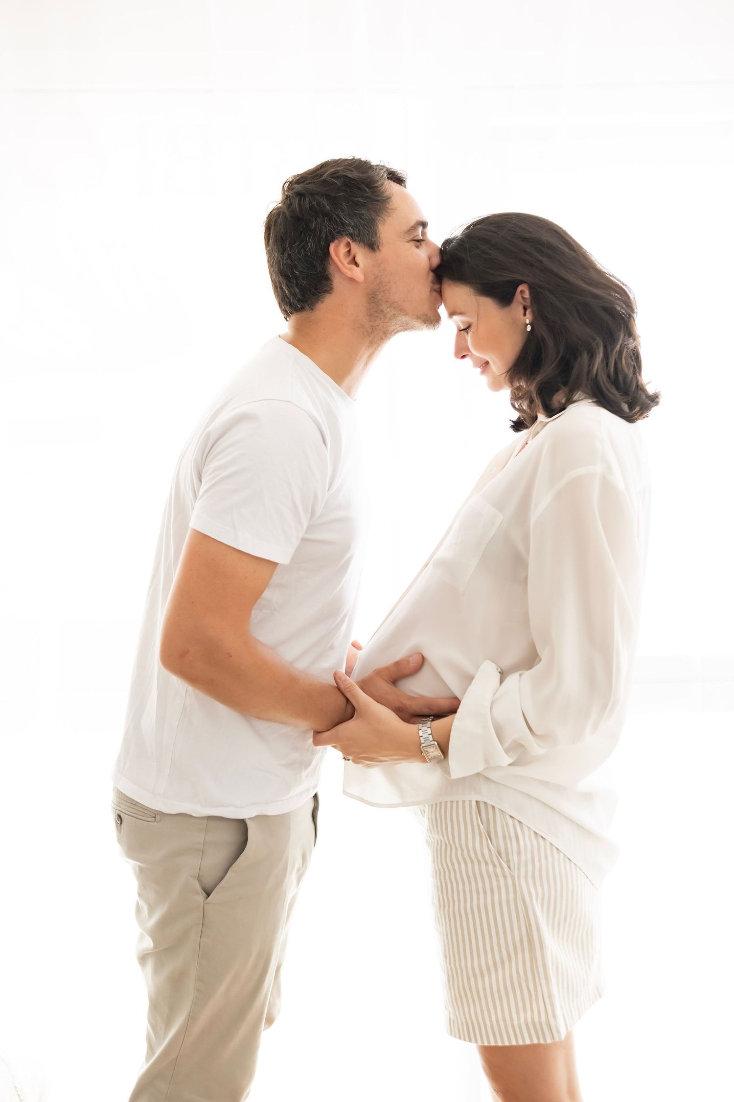 A man kisses a smiling pregnant woman on the forehead while they both cradle her baby bump against a white background.