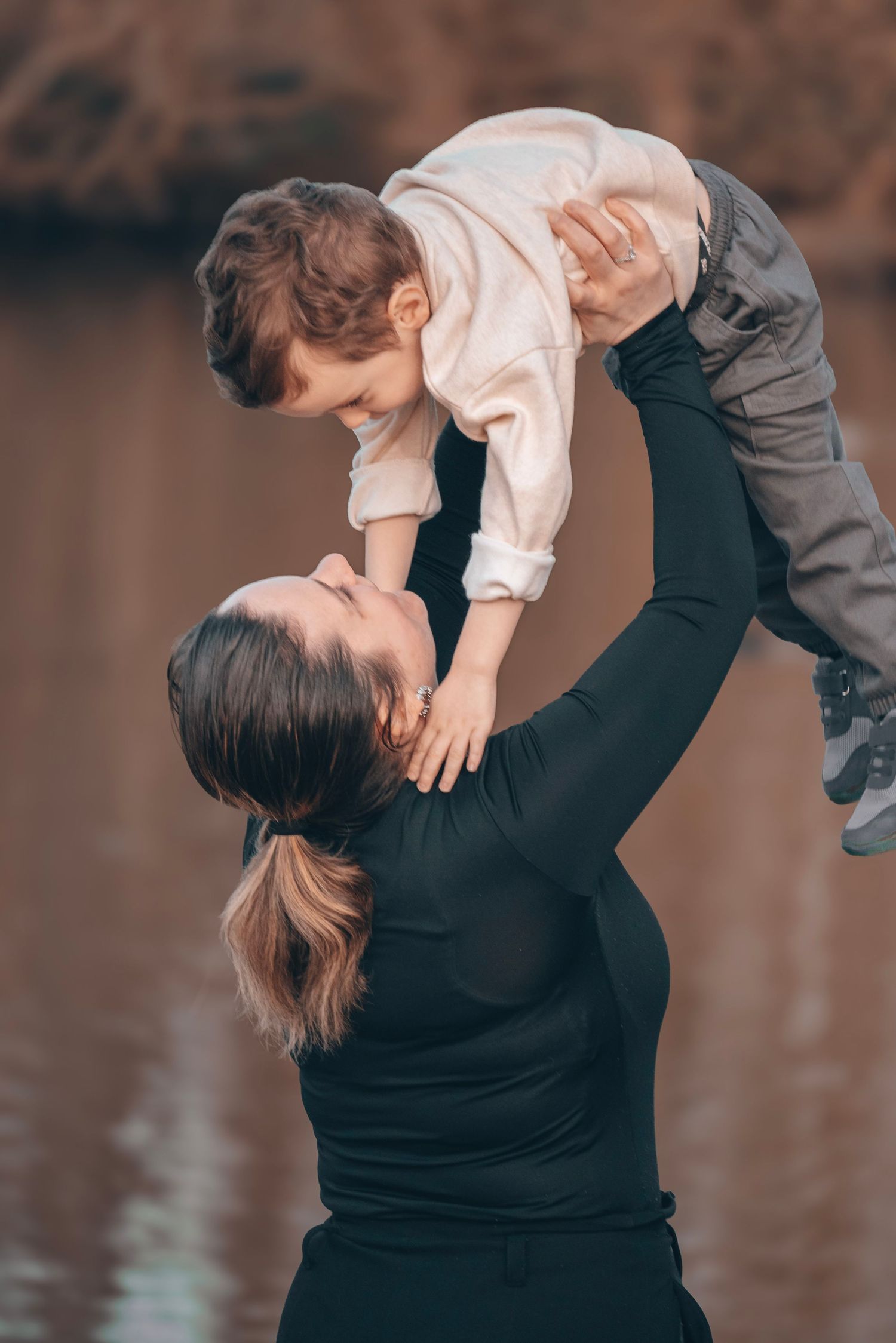 Parent lifts smiling child in air during outdoor photoshoot by the water.