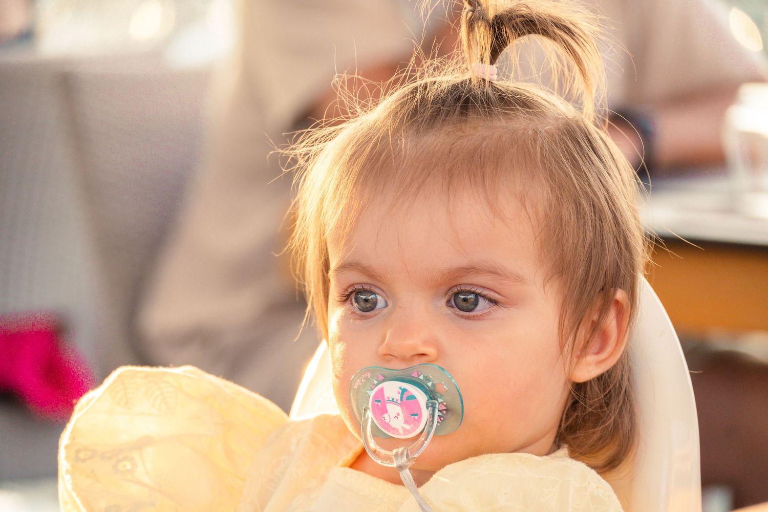 Young child with blonde hair sitting at table with pacifier and holding something yellow in soft golden lighting.