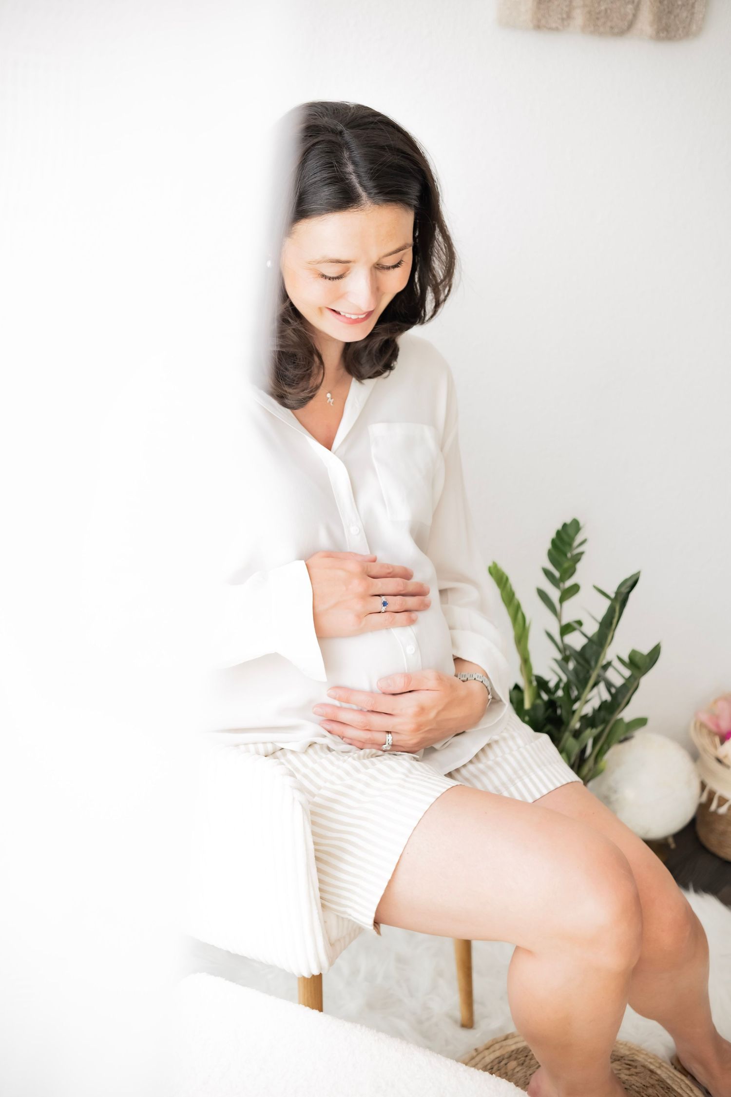 Smiling pregnant woman in white blouse sits on chair cradling baby bump, surrounded by greenery in bright room.