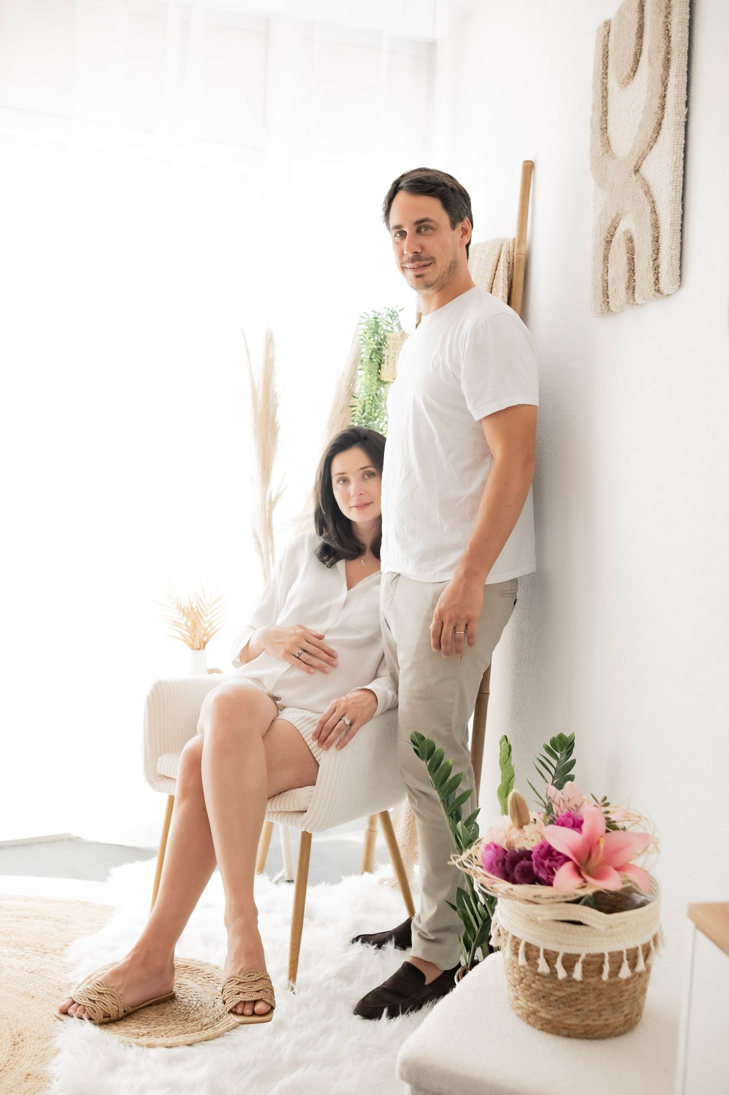 A pregnant woman sits in a chair while her partner stands beside her in a bright, decorated room with flowers.