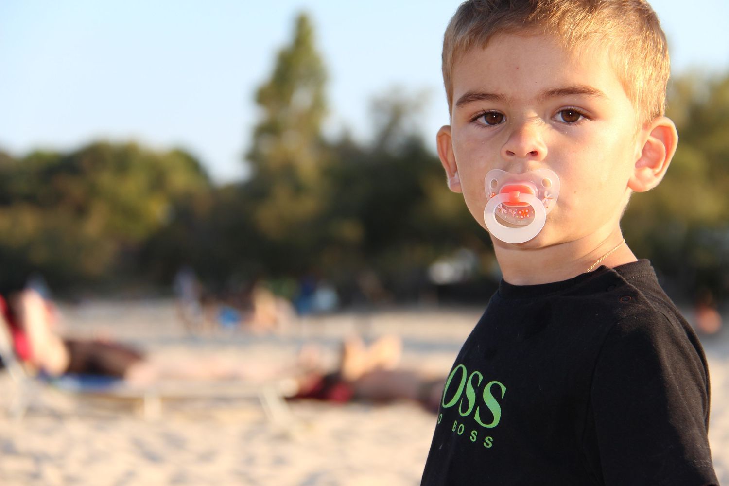A young child in a black t-shirt blows a bubble with bubblegum at the beach during sunset.