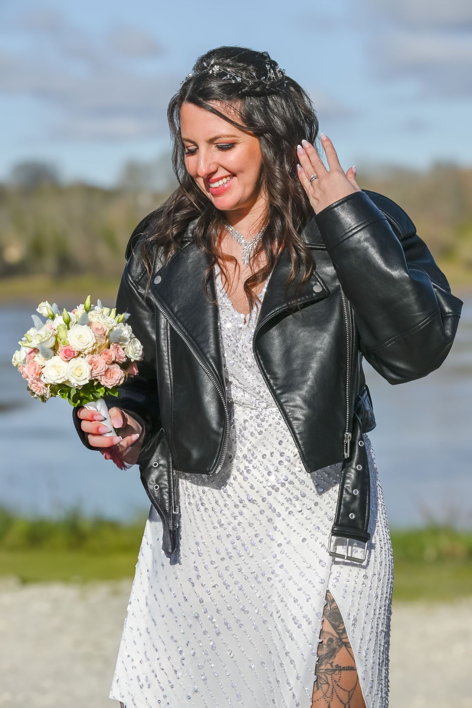 A bride in a white dress and leather jacket holds a pink bouquet by a waterfront.