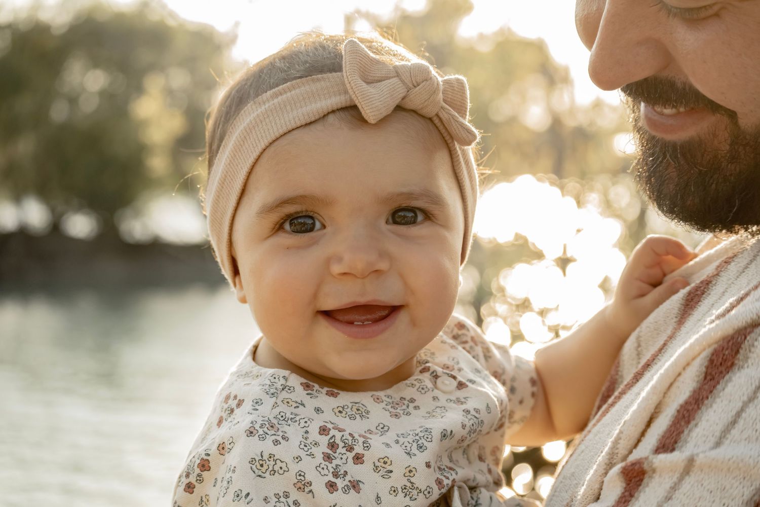 Smiling baby girl with bow headband held by bearded father outdoors in golden sunlight near water.