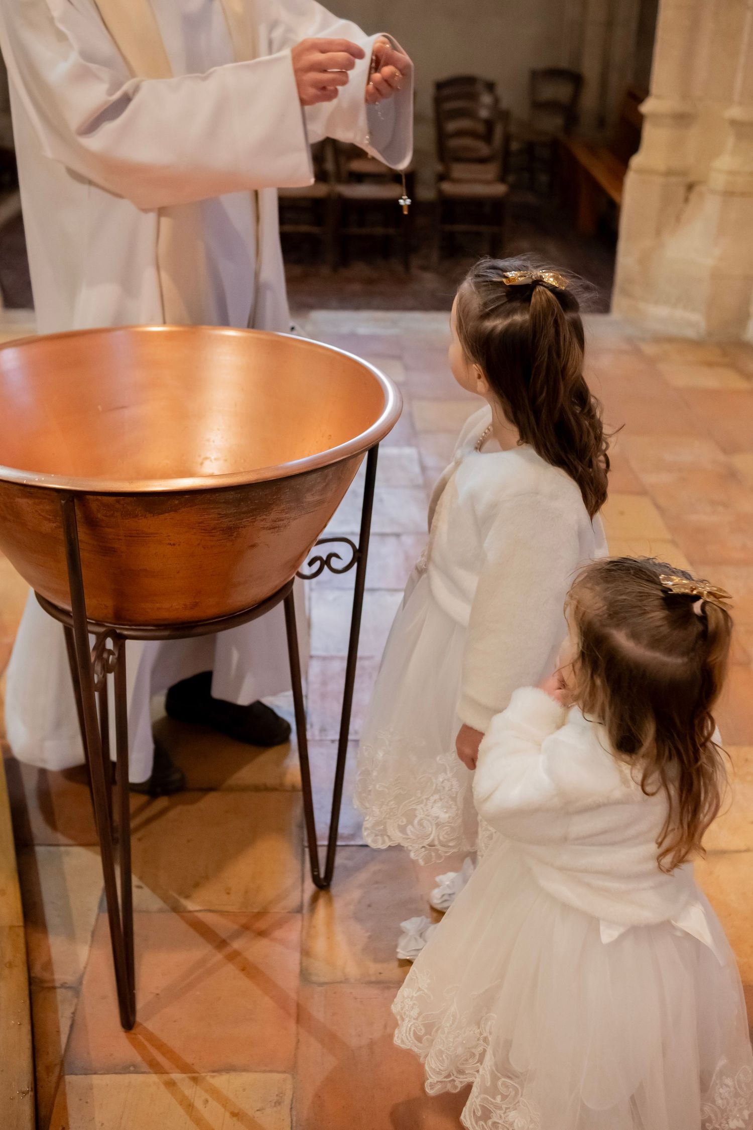 Two girls in white dresses stand by a copper baptismal font during a church baptism ceremony with a priest.