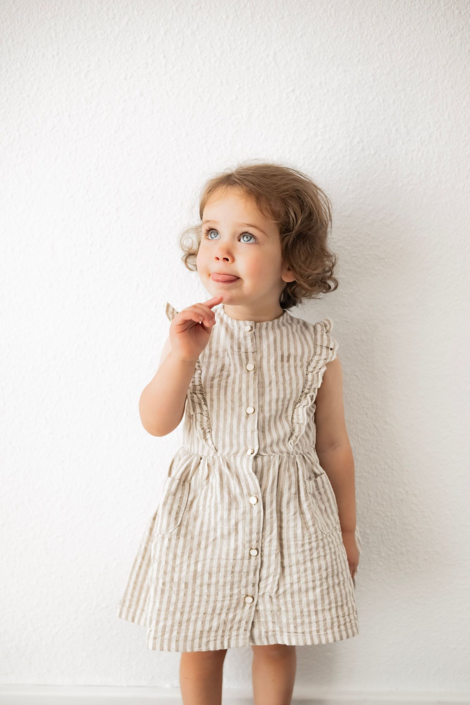 Toddler girl with blue eyes wearing a striped linen ruffle dress, posing against a white wall.