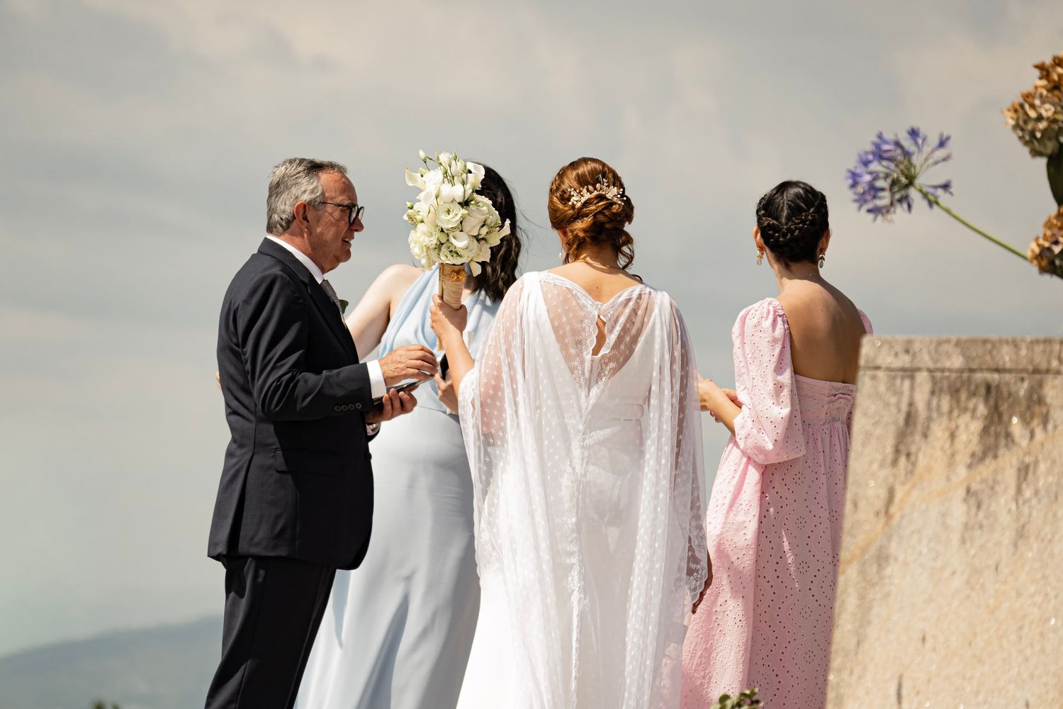Bride in white cape gown with bridesmaids and suited man holding bouquet outdoors at scenic wedding venue.