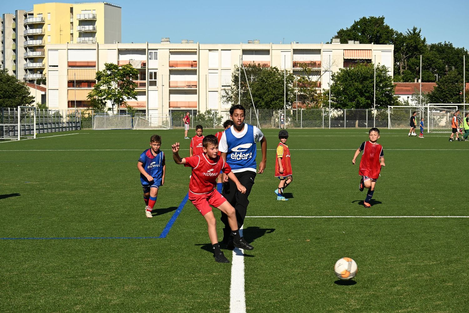 Young soccer players competing in a match on a green field with apartment buildings visible in the background.