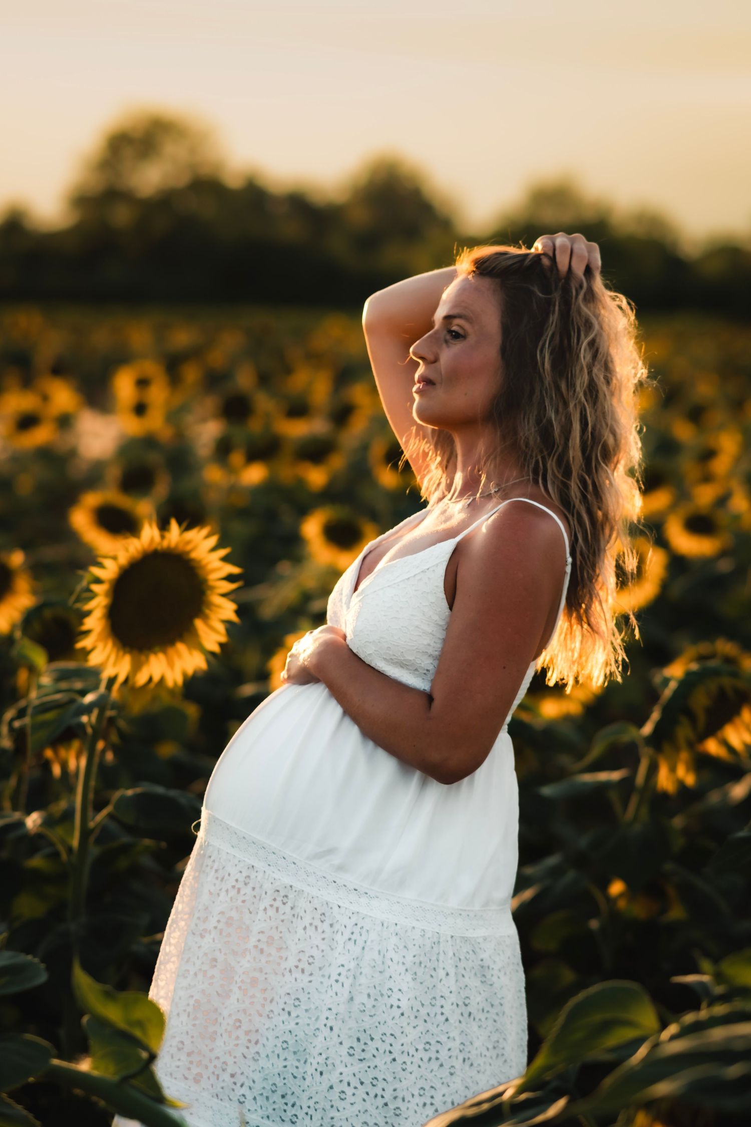Pregnant woman in white dress standing in sunflower field at golden sunset hour.
