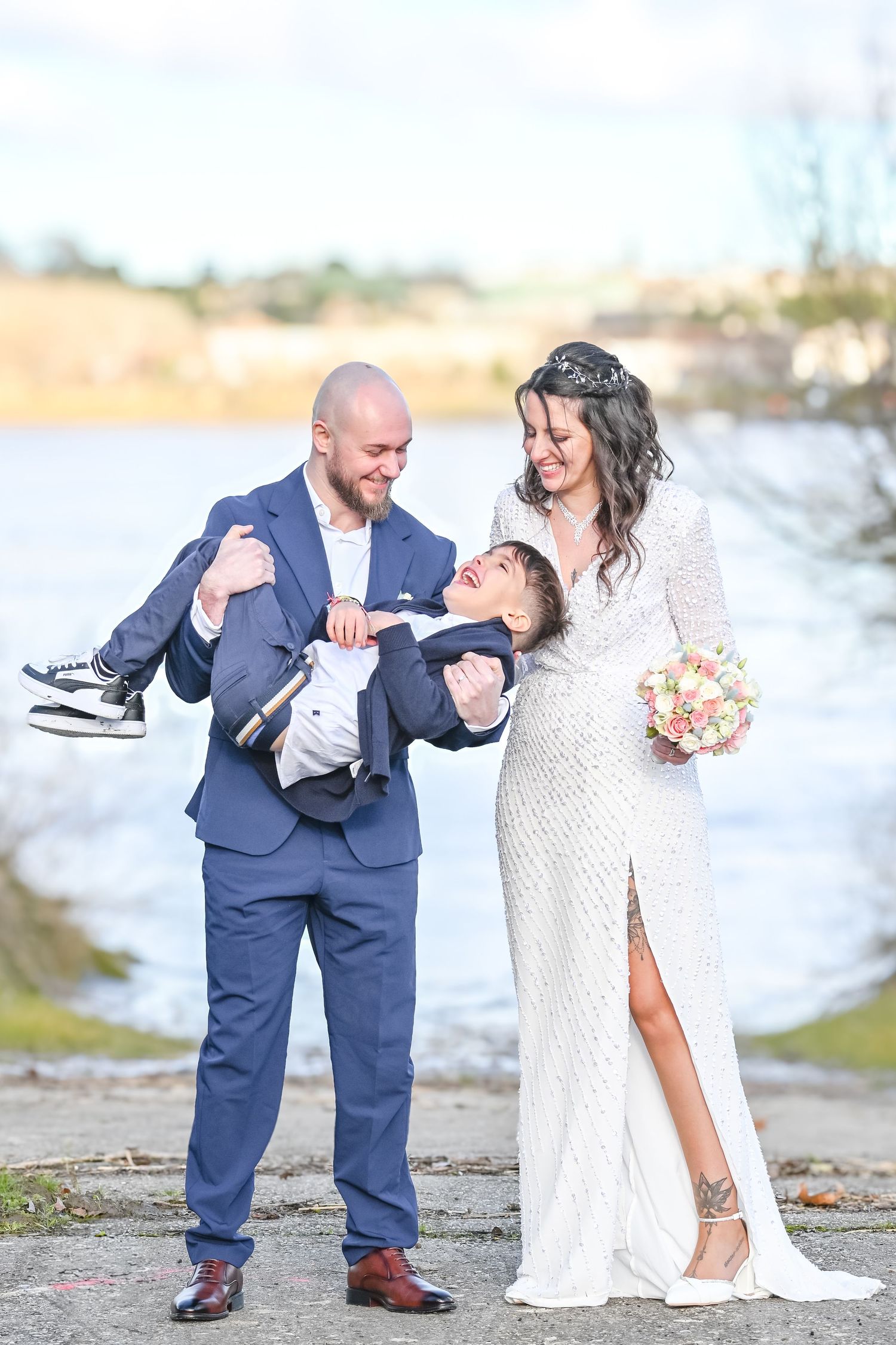 Wedding couple in elegant attire pose by water with their little one.