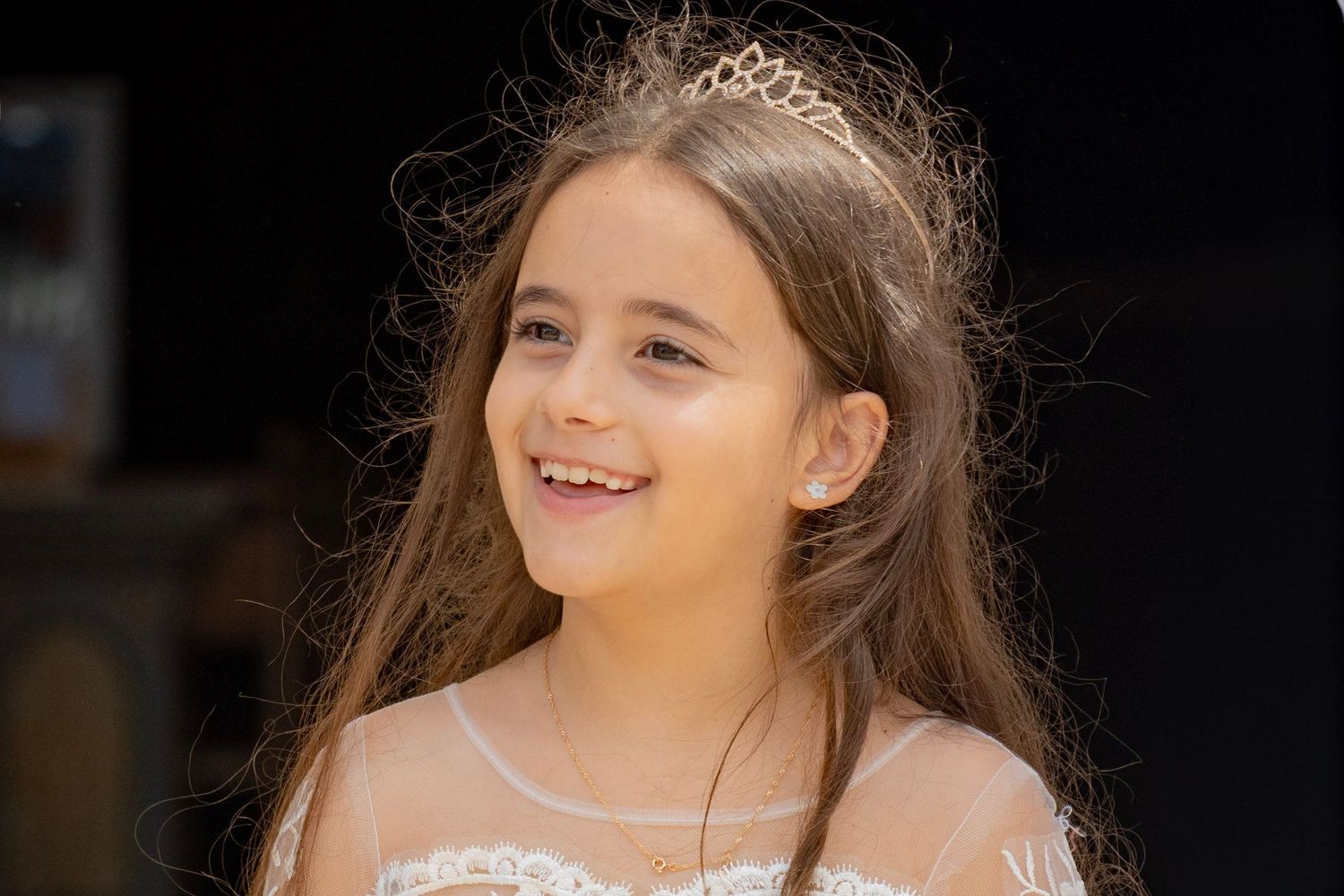 Young girl smiling in white lace dress with gold tiara, long brown hair, joyful expression at outdoor event.