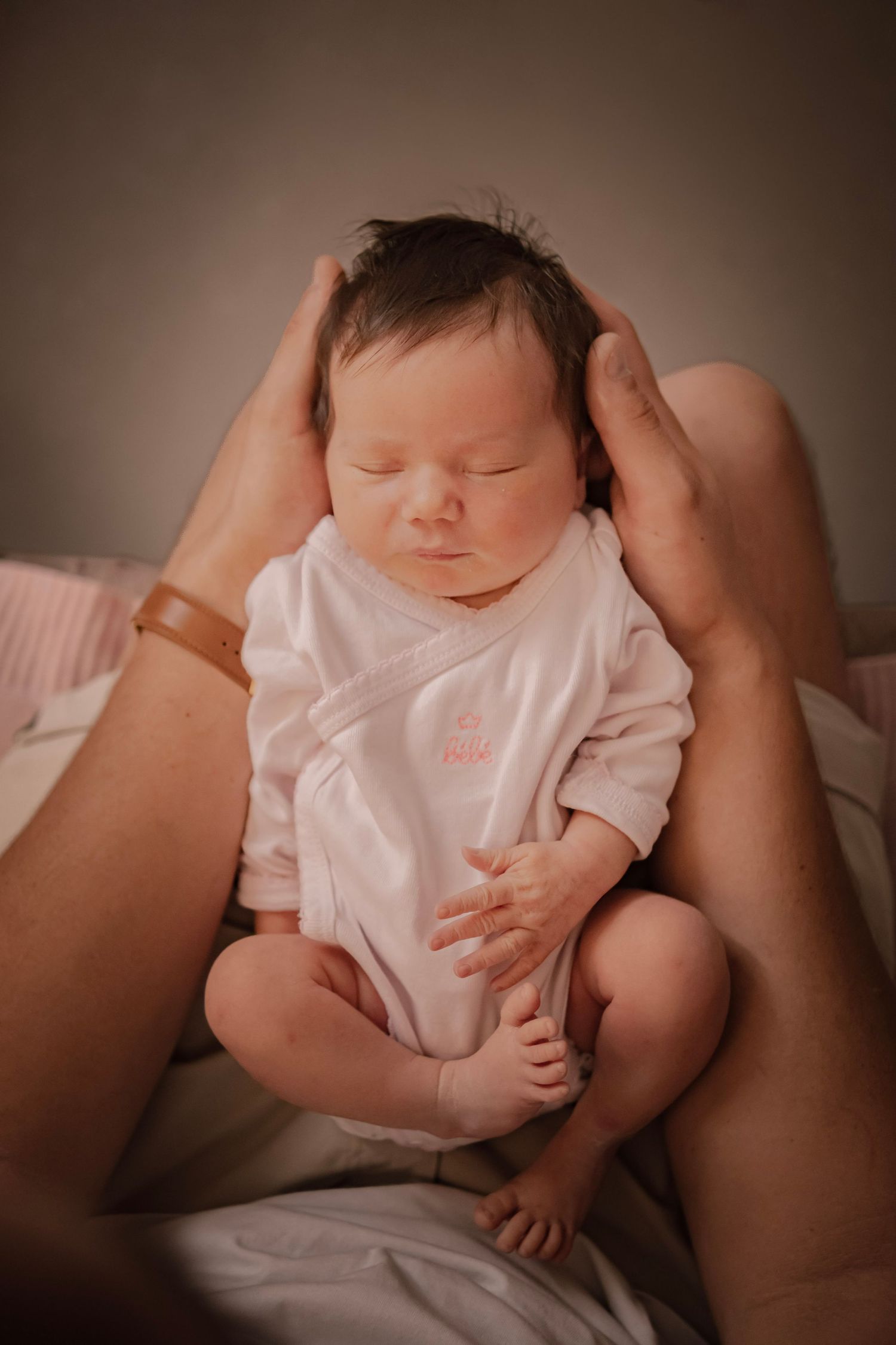 Newborn baby in pink onesie sleeping peacefully while being cradled in parent's hands on a soft bed.