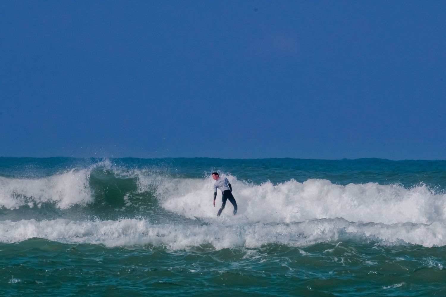 A surfer rides an ocean wave against a bright blue sky in turquoise waters.