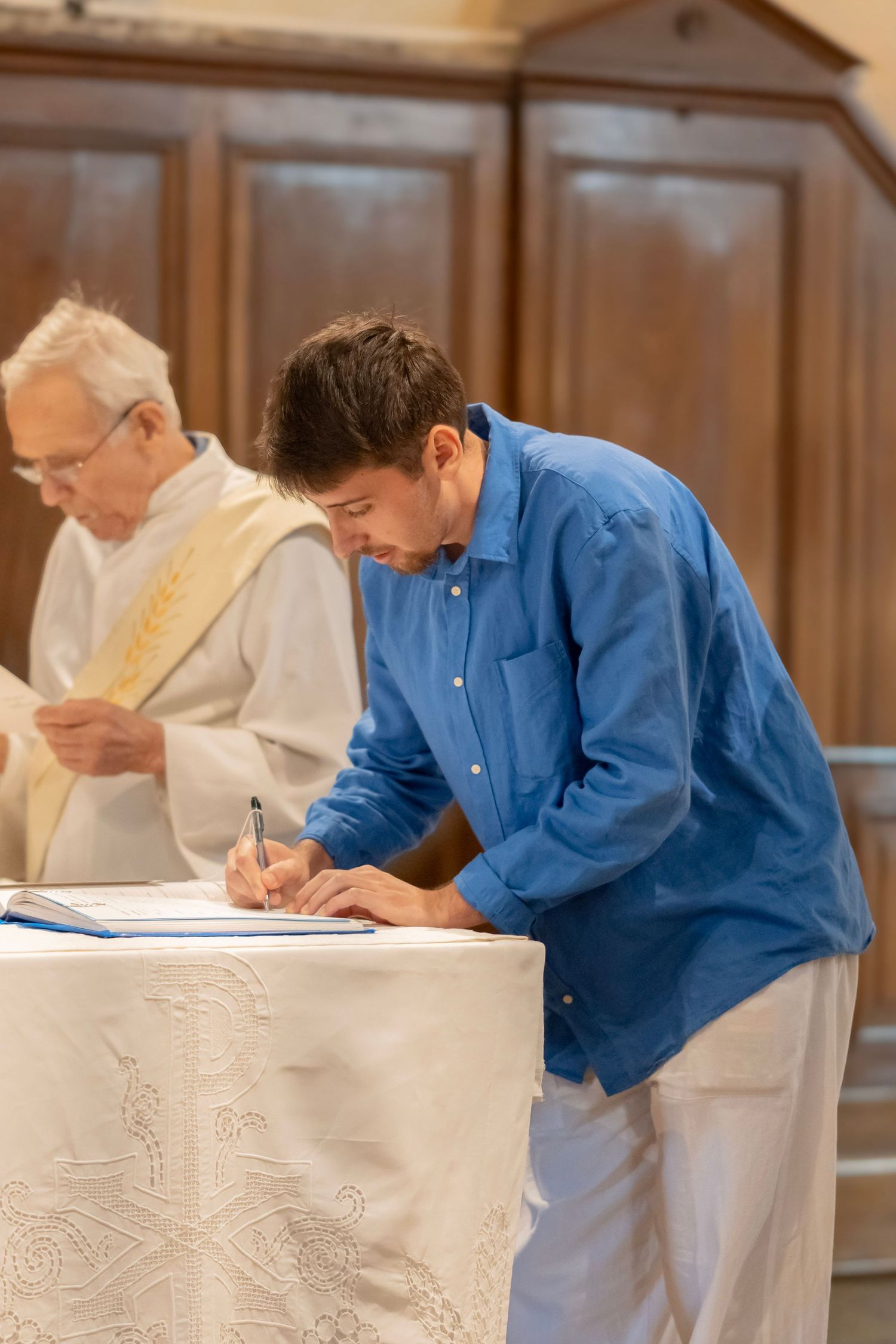 Two people in blue and white clothing lean over a table while writing or signing documents in an indoor setting.