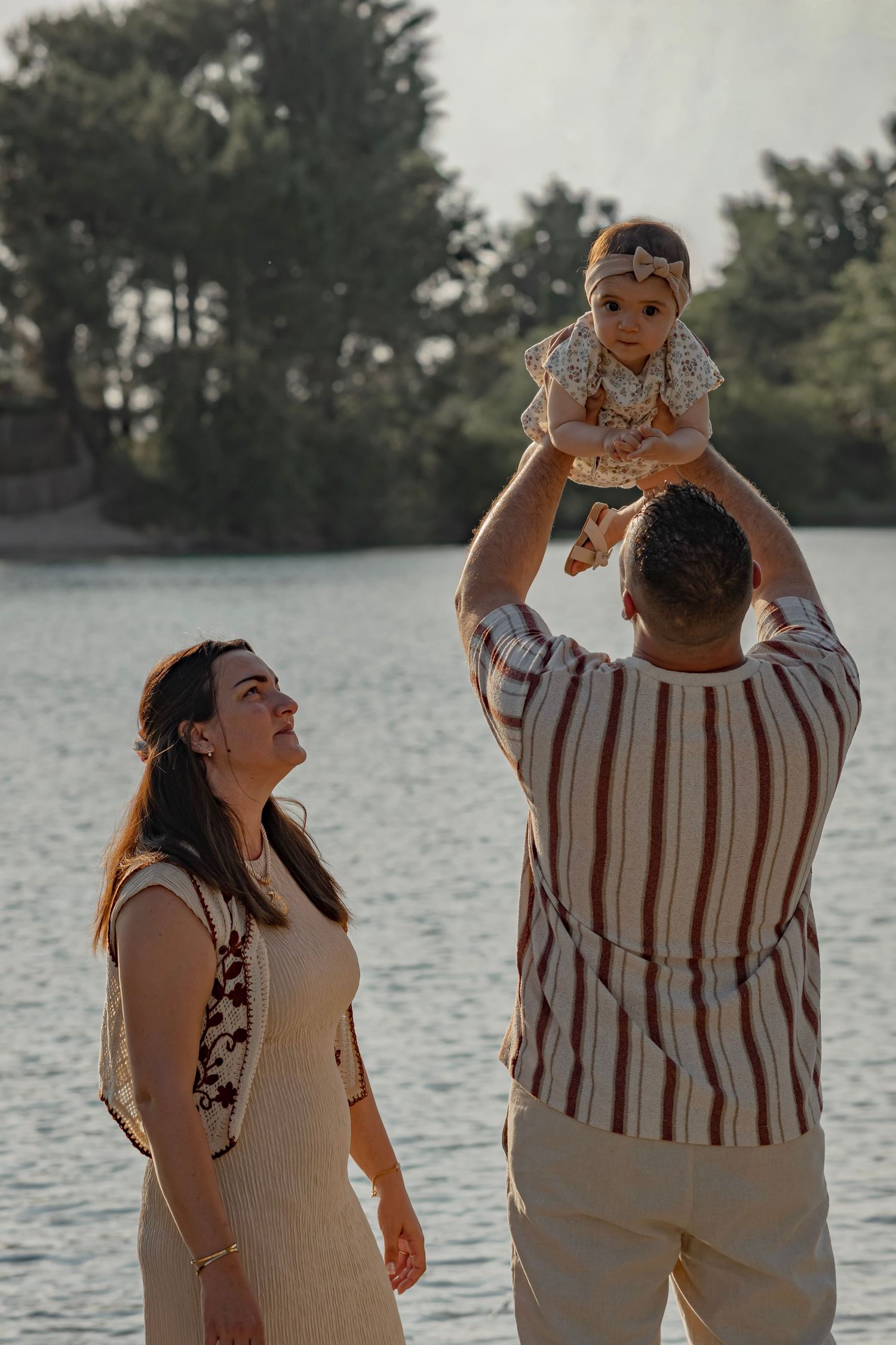 A couple enjoys a sunny lakeside moment, with the father lifting their baby girl overhead while the mother watches and smiles.