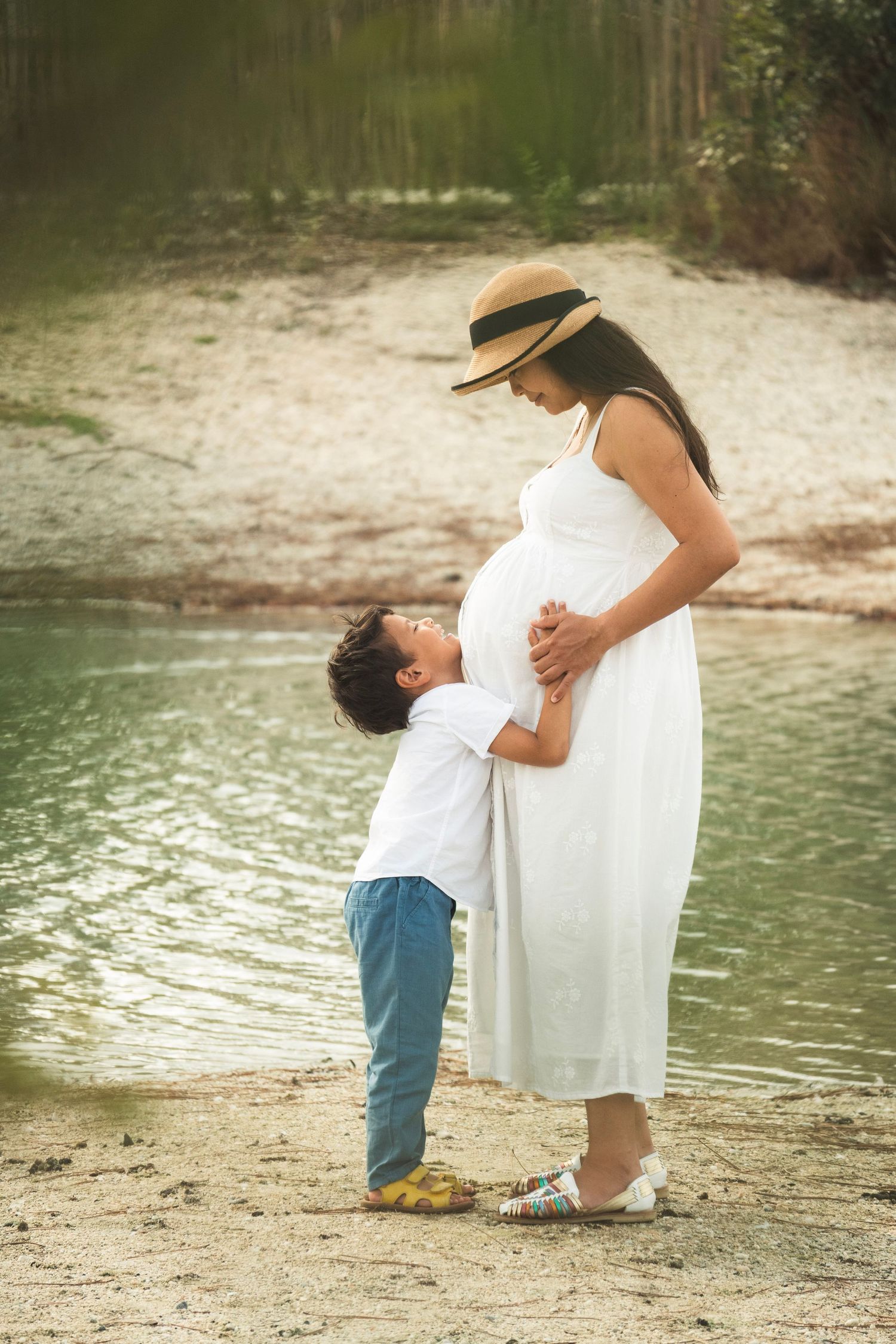 A young boy kisses his pregnant mother's belly near a serene lake, as she wears a white dress and straw hat.