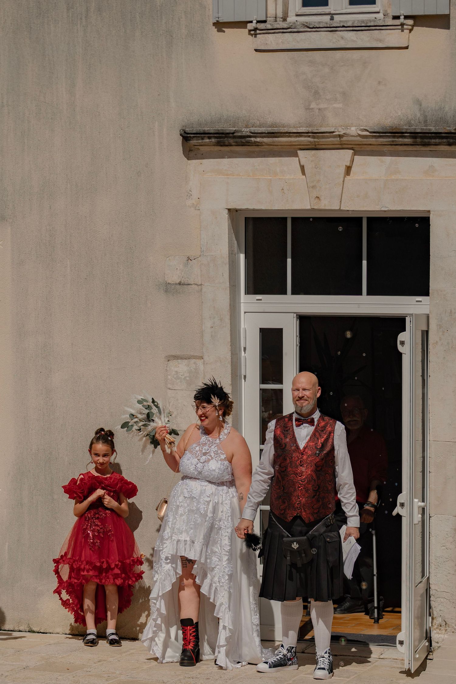 Bride in white dress and groom in kilt exit building with flower girl in red dress at outdoor wedding ceremony.