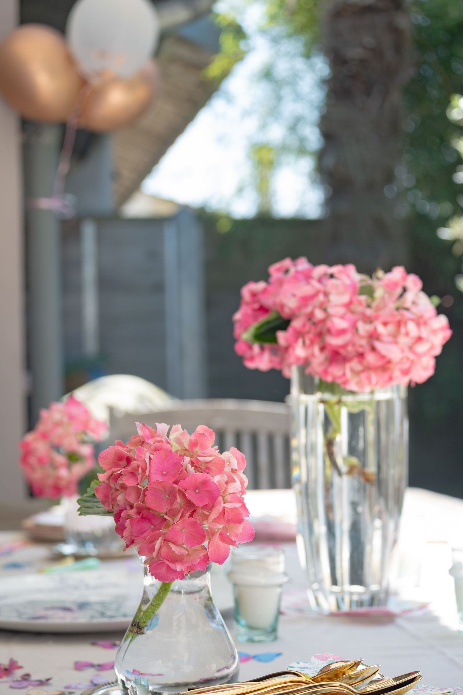 Pink hydrangeas in glass vases decorate an outdoor party table with gold cutlery and balloons in the background.