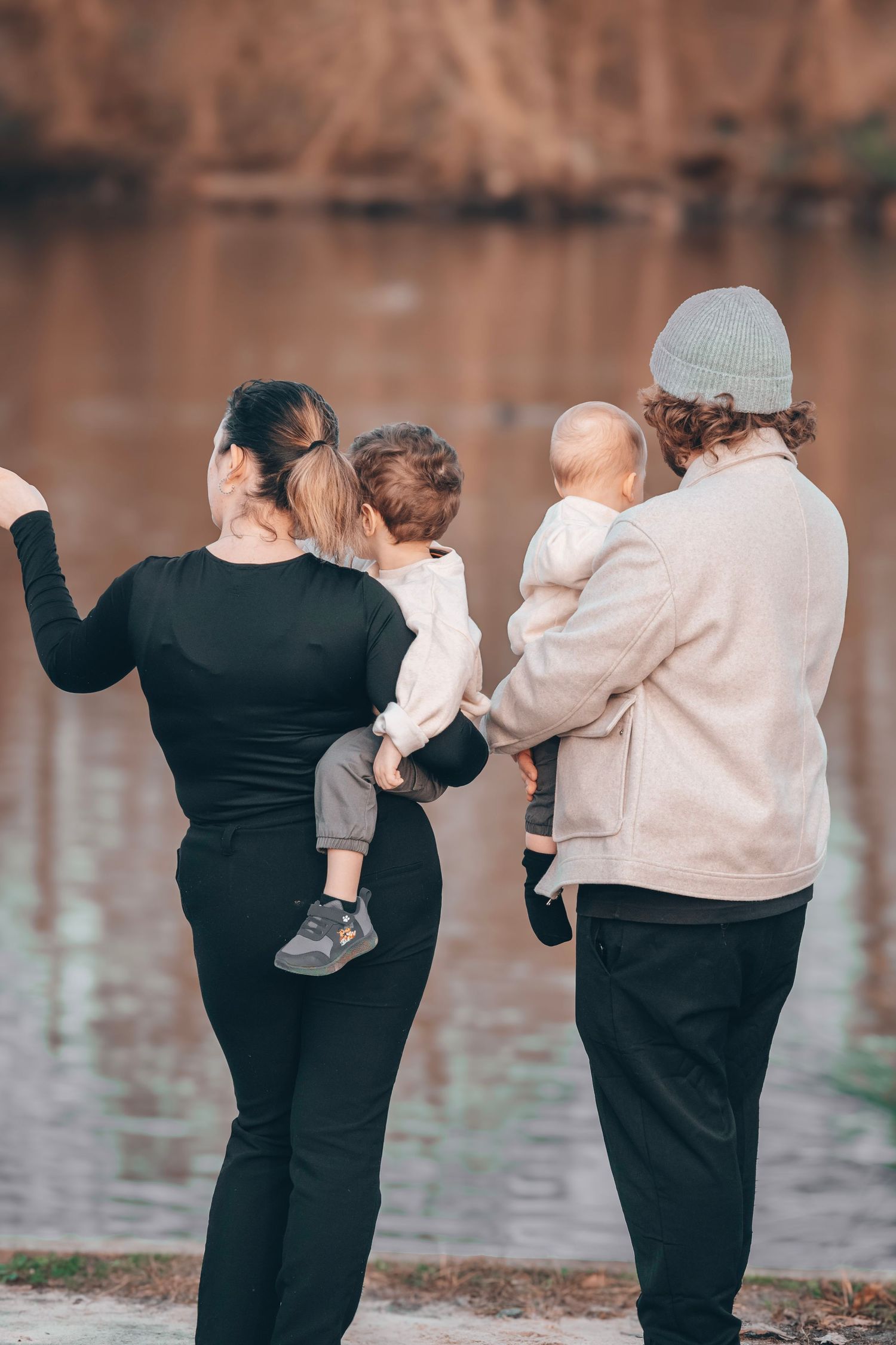 Two adults hold small children while standing by a lake at dusk.