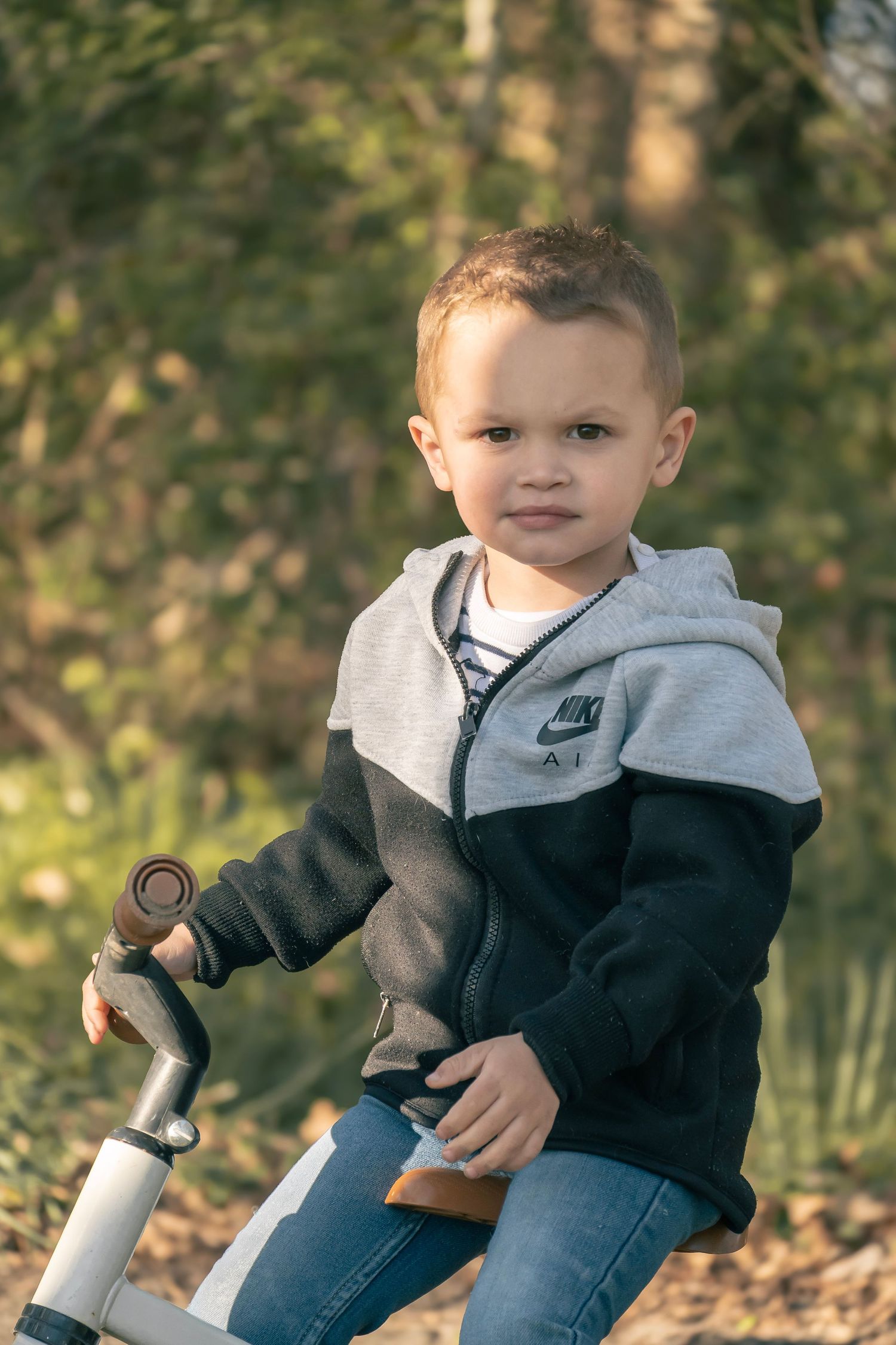 Small child in a Nike hoodie sits on a bike outdoors during autumn with trees in the background.