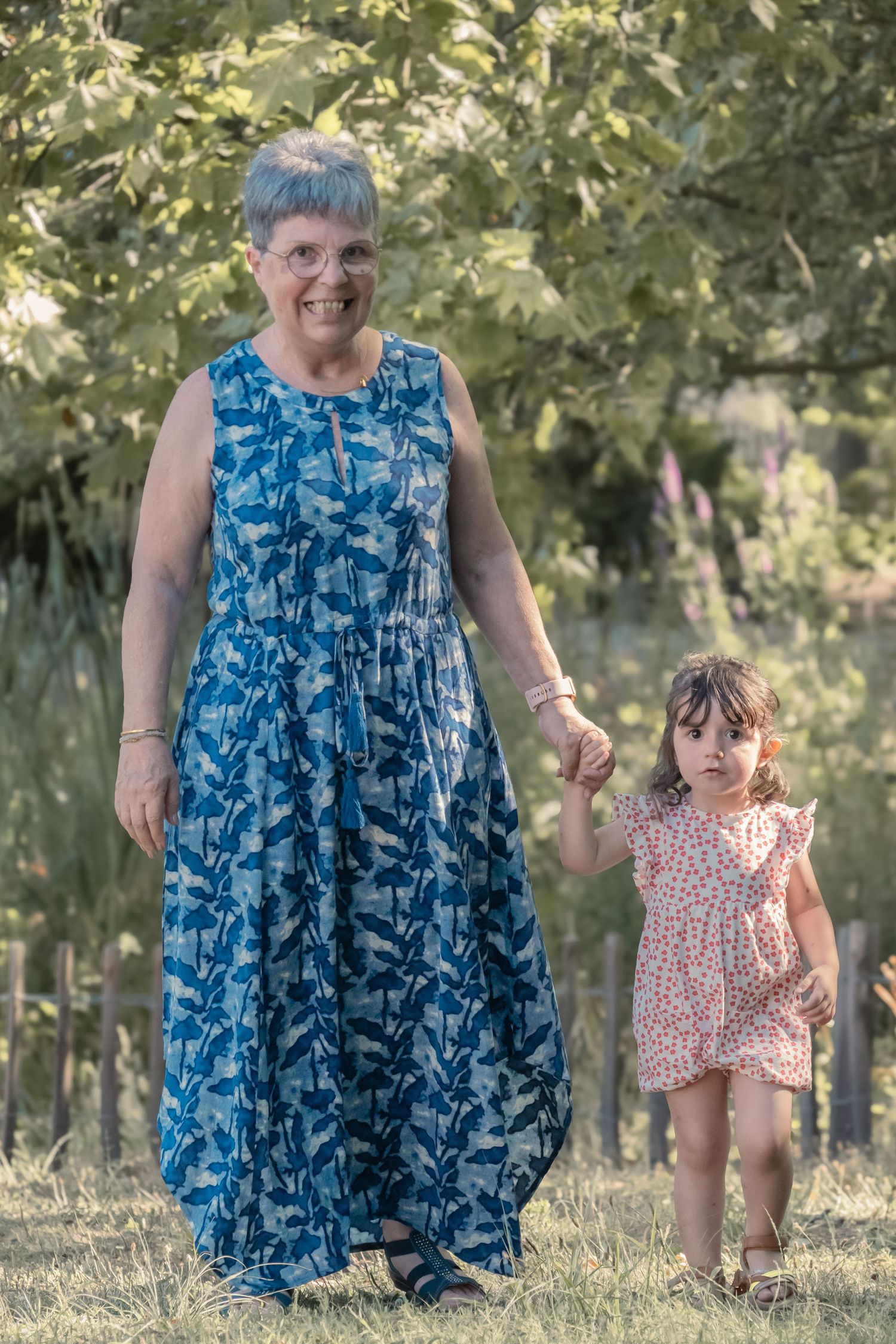 A pair in summery dresses hold hands while standing in a grassy outdoor setting with trees in the background.