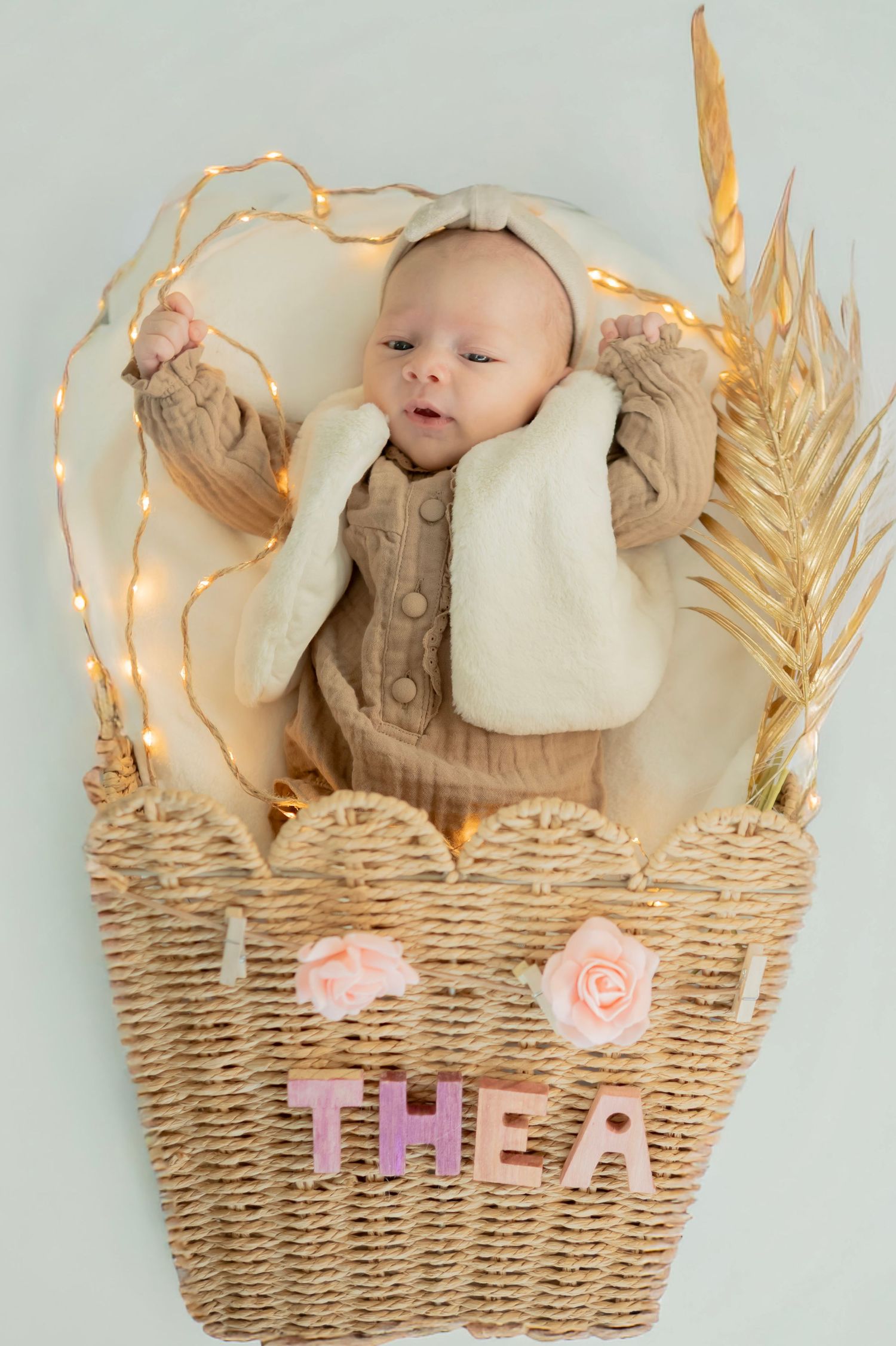 A cozy woven basket decorated with pink roses and string lights holds a small baby in warm neutral tones.
