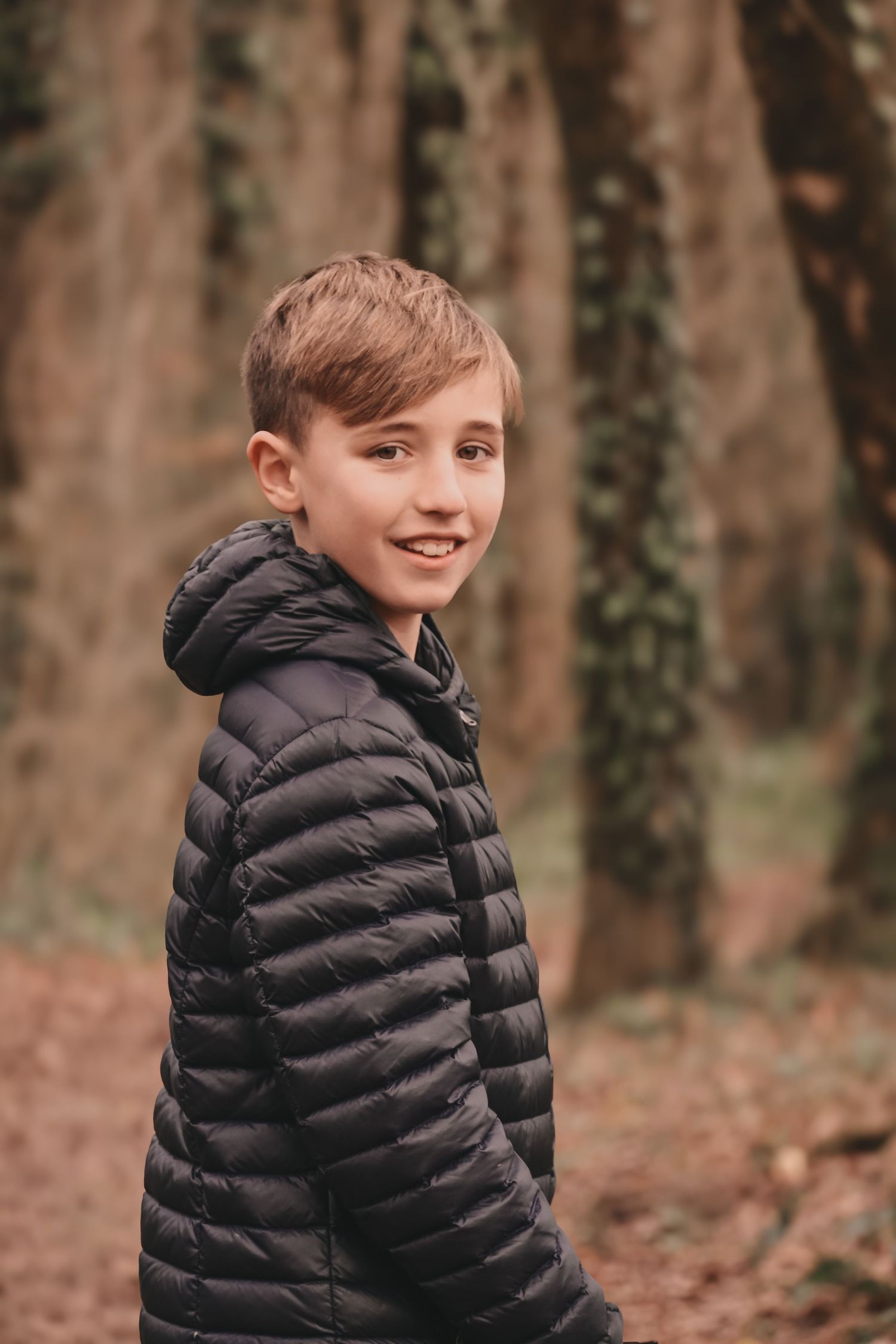 Child in a black puffer jacket standing in a forest setting.