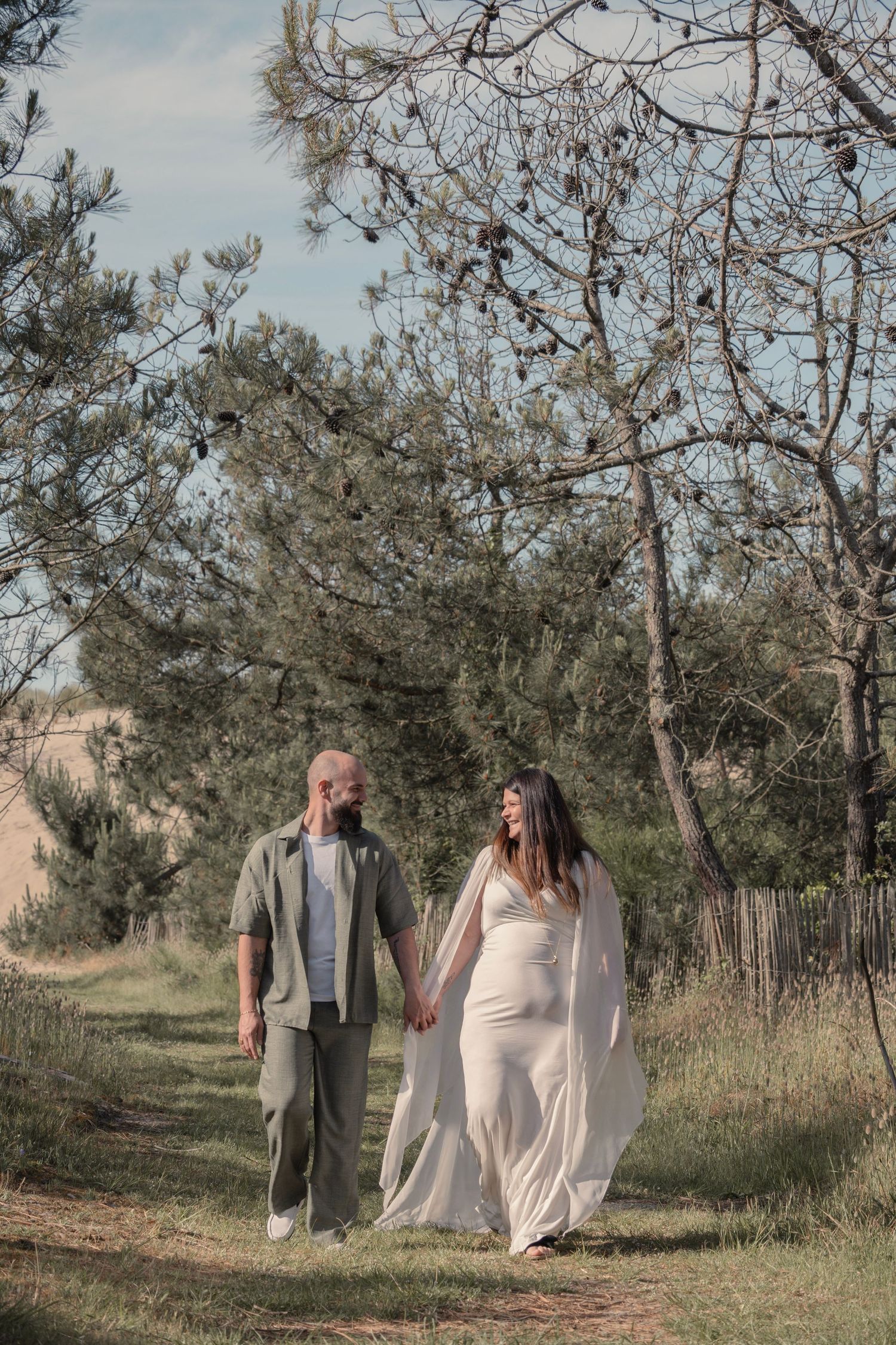 A couple walks hand in hand along a rustic path bordered by pine trees on a tranquil autumn day.